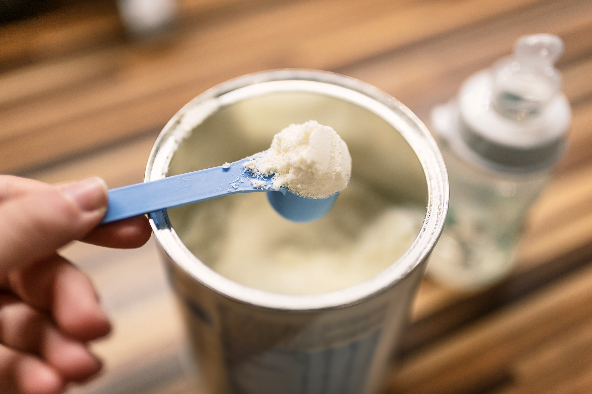 Hand holding small measuring blue scoop of baby formula over canister of it, selective focus, baby bottle on the right, lined wooden table blurred in the background