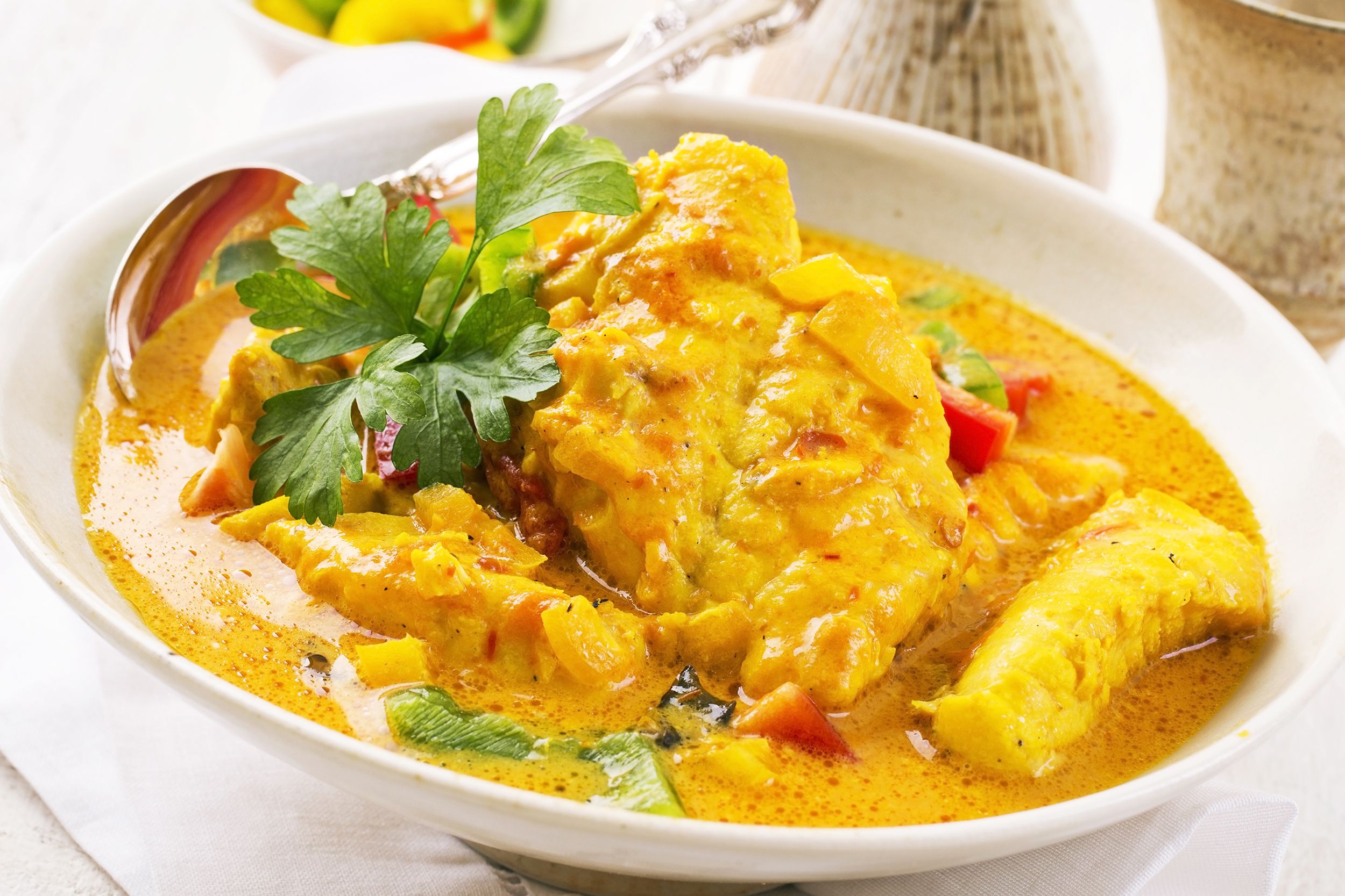 Thai red curry fish stew in a white porcelain bowl with a silver spoon on the left side, on a white napkin, three serving vessels blurred in the background