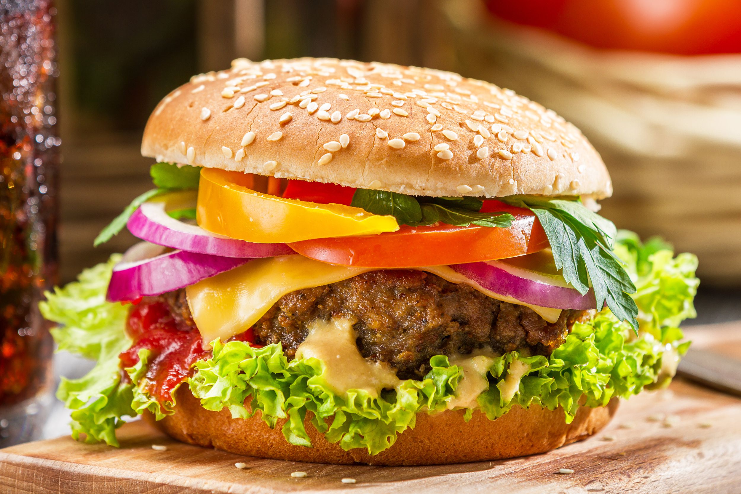 The best burger, stacked with fresh lettuce, onions, tomatoes, and peppers, on a wooden cutting board, with a glass of cola to the left and a blurred background