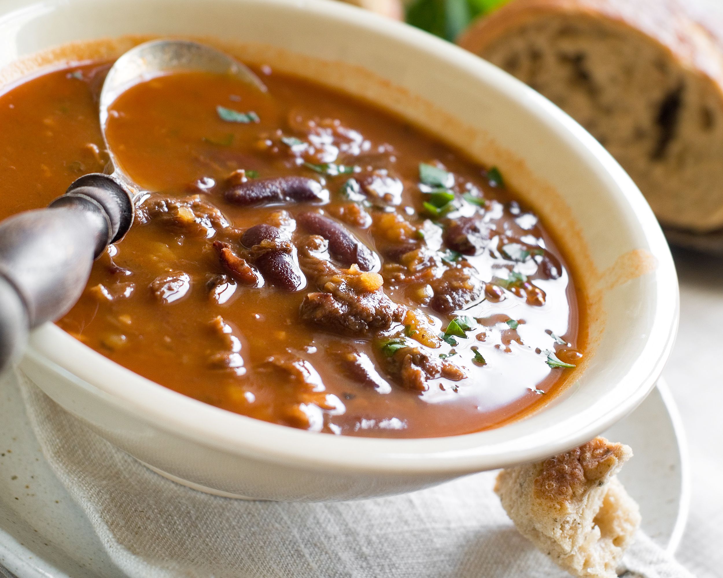 Three-bean chili with a spoon in a white bowl, diagonal, on a beige napkin with a piece of bread, on a brown speckled white bowl with a blurred background of bread and a white table