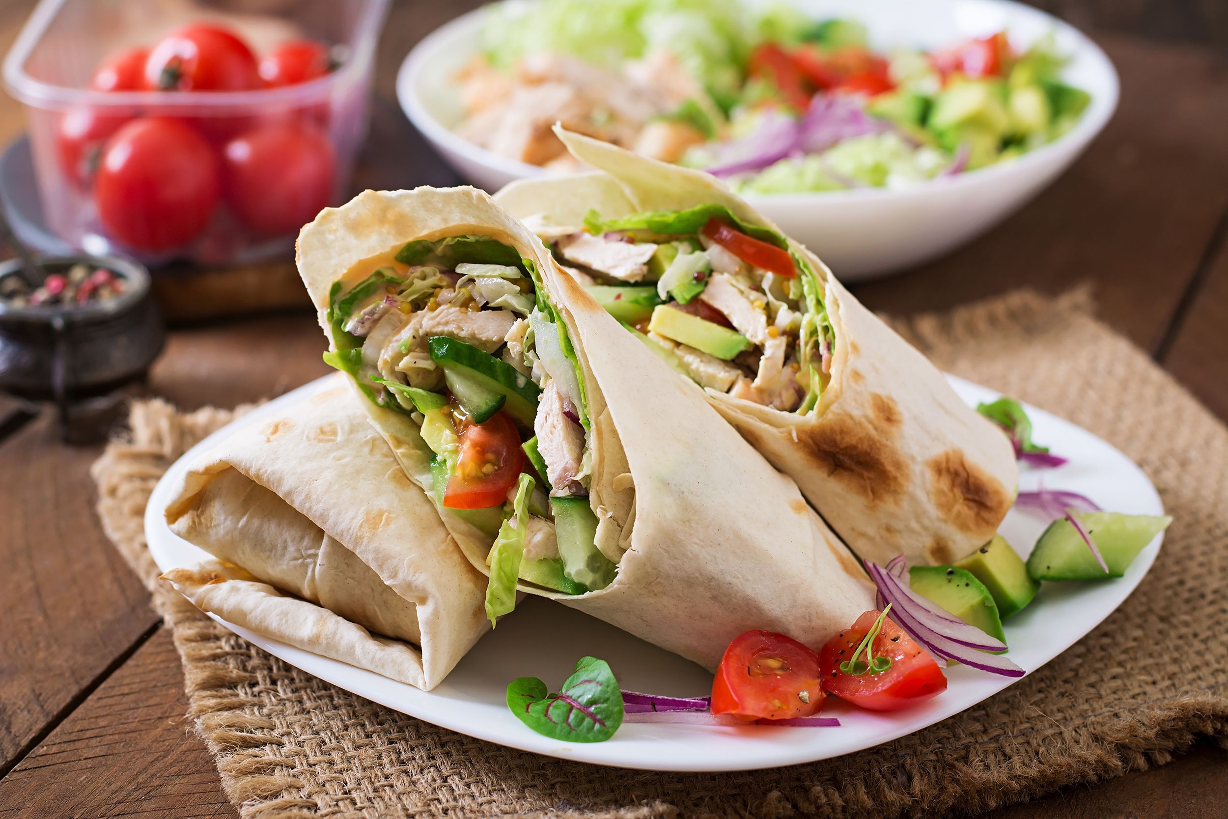 Three crispy chicken and avocado wraps on a white plate on a rustic napkin and wooden table with a blurred background of a bowl of salad and tomatoes