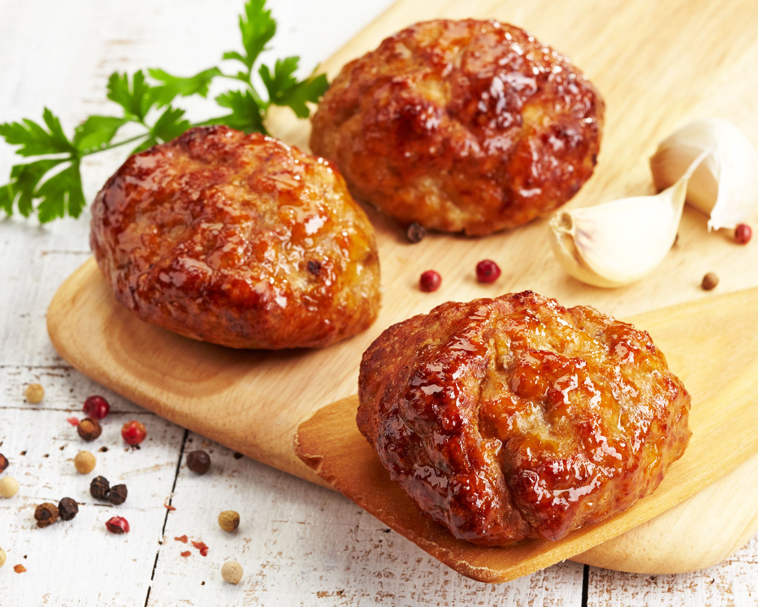 Three large baked turkey meatballs, one is on a wooden spatula while the other two is on a wooden serving board on a white wooden table with peppercorns scattered