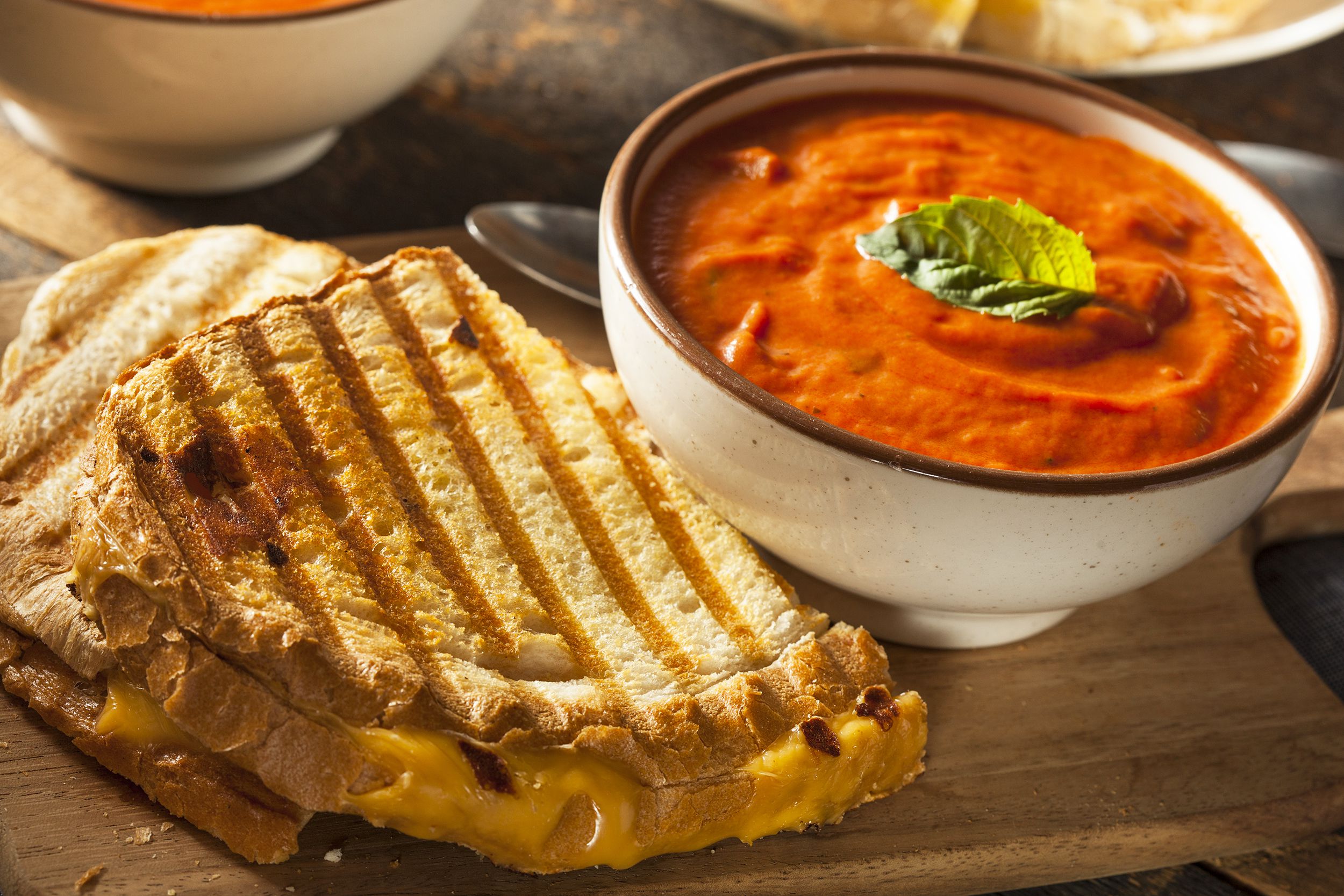 Tomato soup in a brown rimmed white ceramic bowl next to two pieces of a grilled-cheese sandwich with a spoon on a wooden serving tray, with another bowl of tomato soup and a rustic table blurred in the background