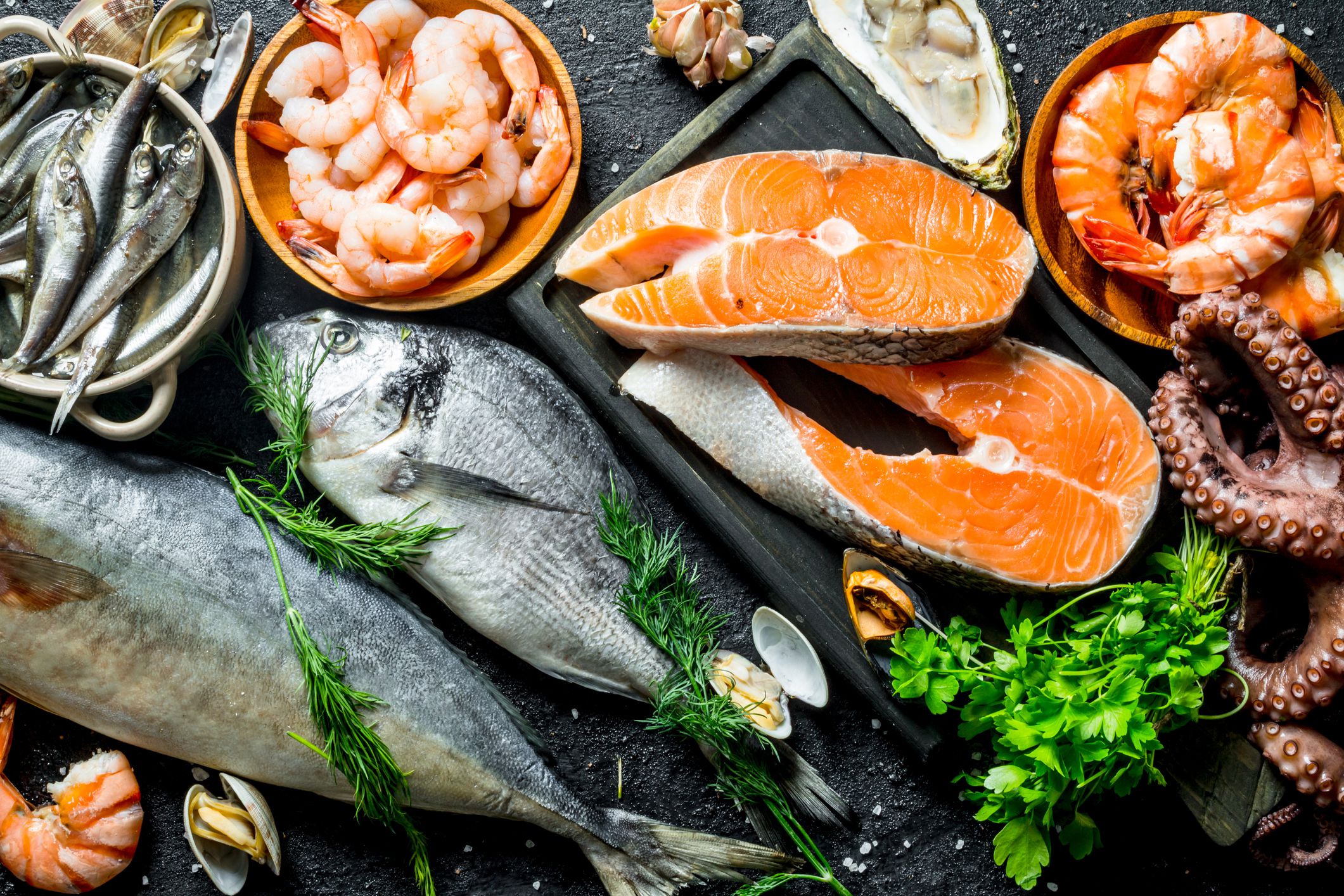 Top view of several different kinds of seafood including slices of salmon, a bowl of shrimp, a bowl of sardines, a bowl of lobster, oysters, octopus, and more with herbs on a large slate board