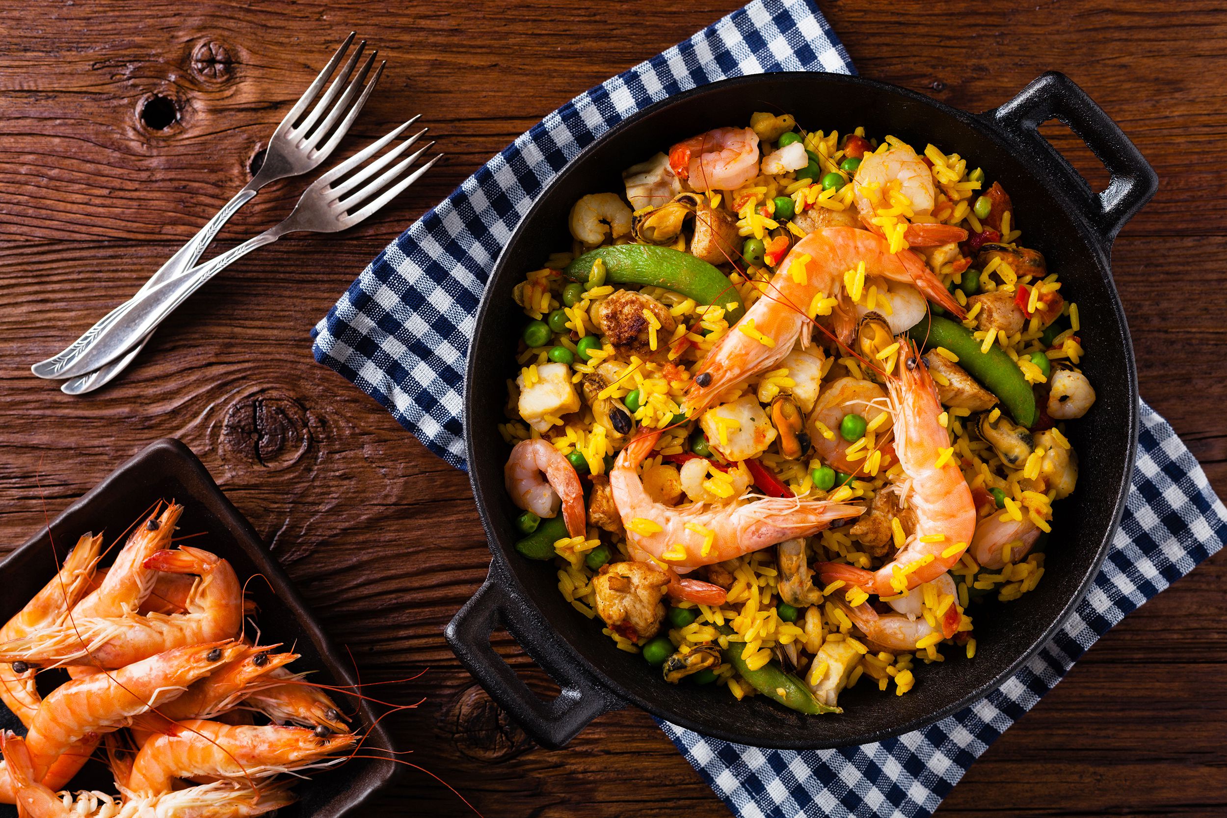 Top view of Spanish shrimp paella in a round cast iron skillet on a blue checkered napkin with a square brown bowl of natural cooked shrimps in the bottom left on a dark brown wooden table with two forks on the top left
