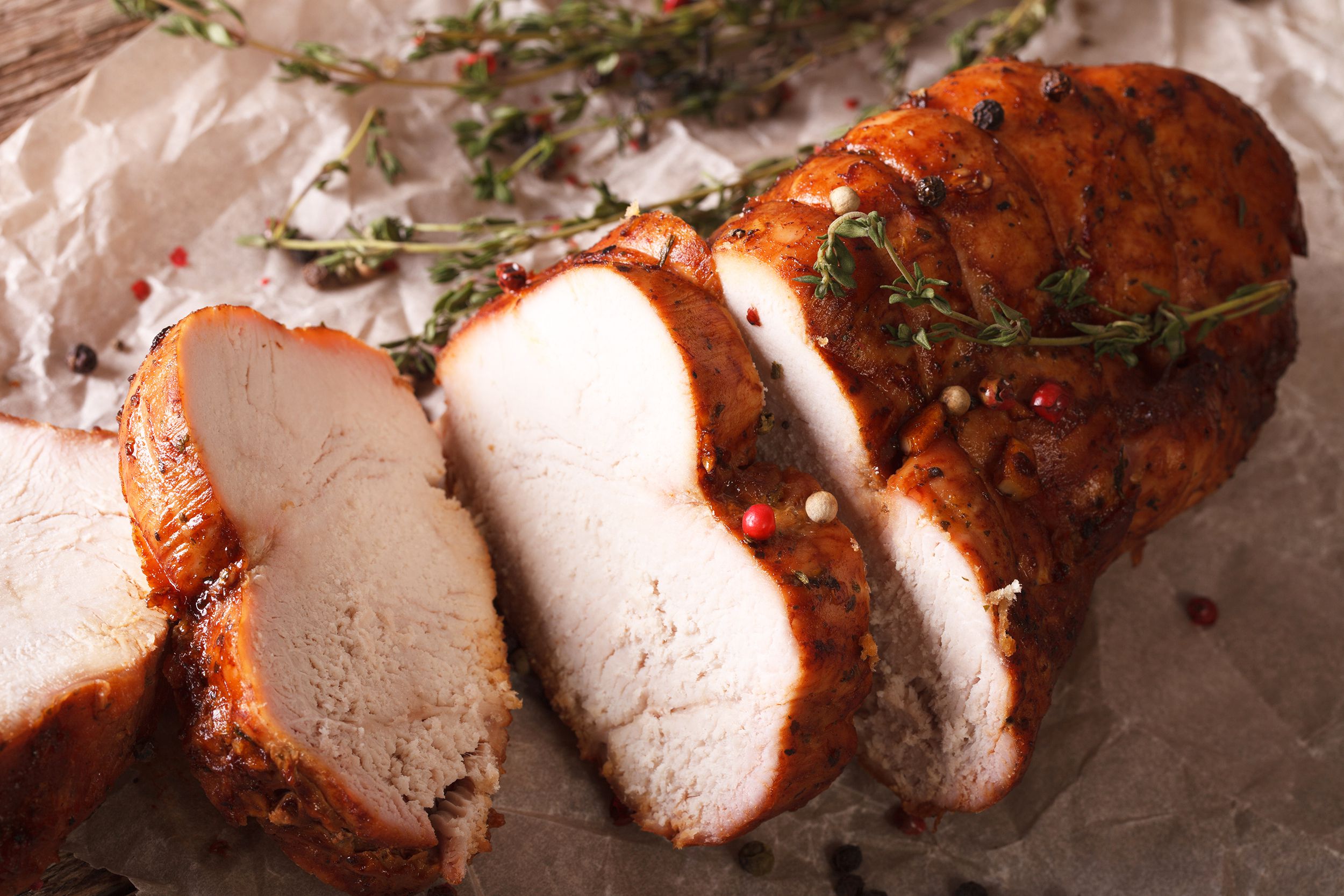 Top view of an oven-roasted turkey breast partially cut into slices on crumbled white paper, surrounded by herbs