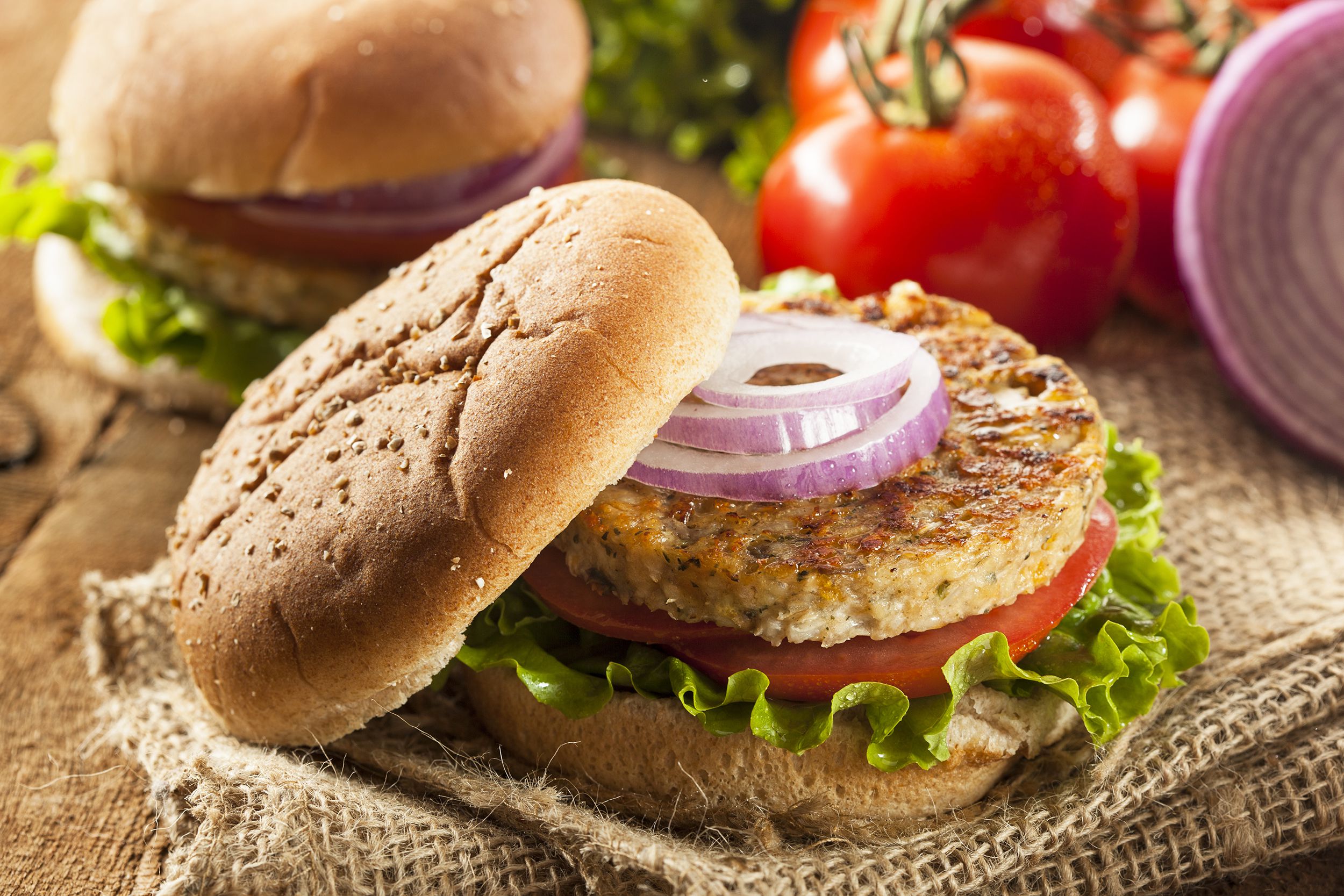 Two easy grillable veggie burgers, one in the center with the top bun off, revealing slices of onion, on a natural napkin surrounded by ingredients blurred in the background