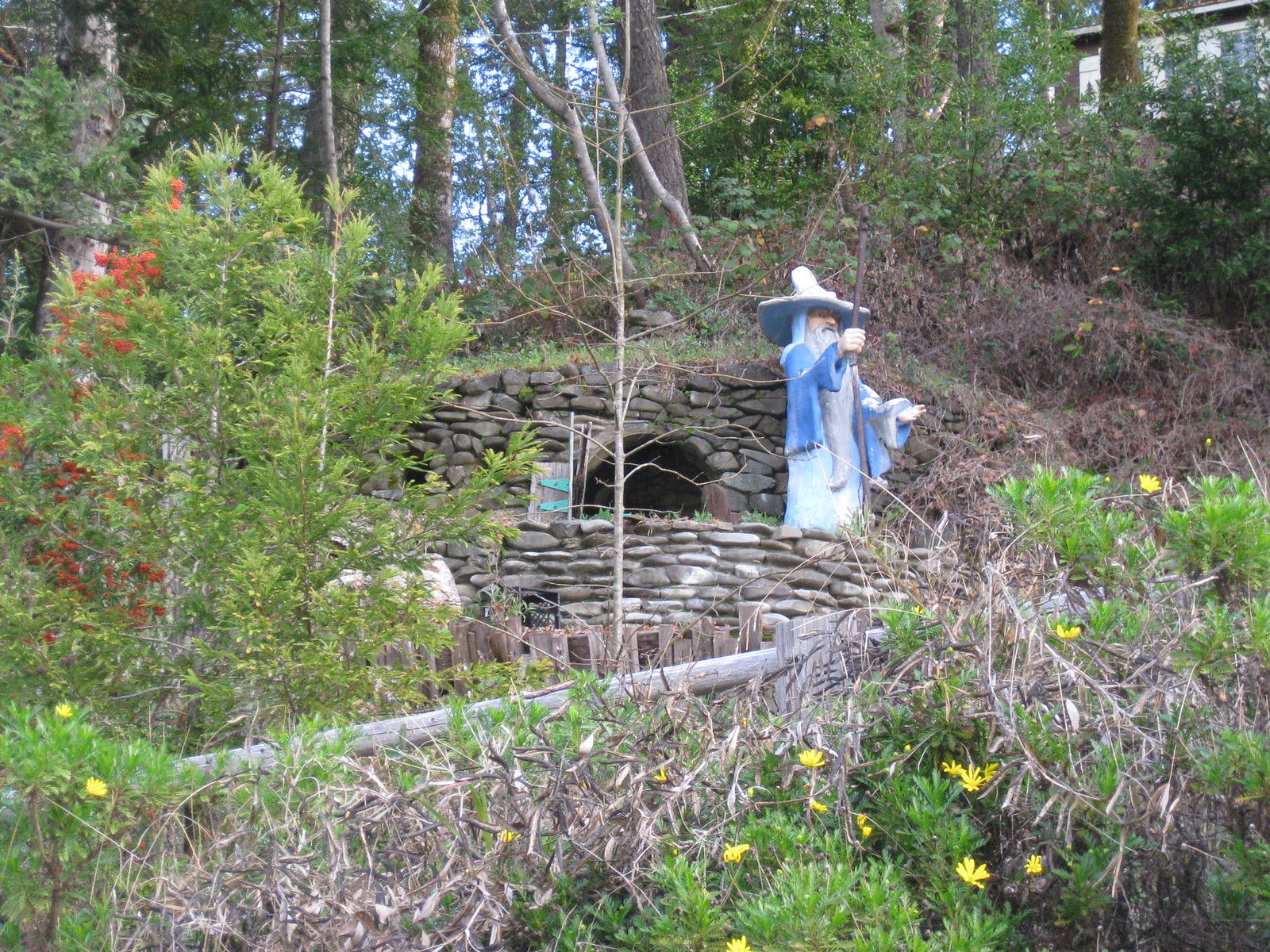 Abandoned Hobbiton USA, Phillipsville, California, hobbit statue on a stone fence, surrounded by plants and trees, trees in the background and a blue sky coming through