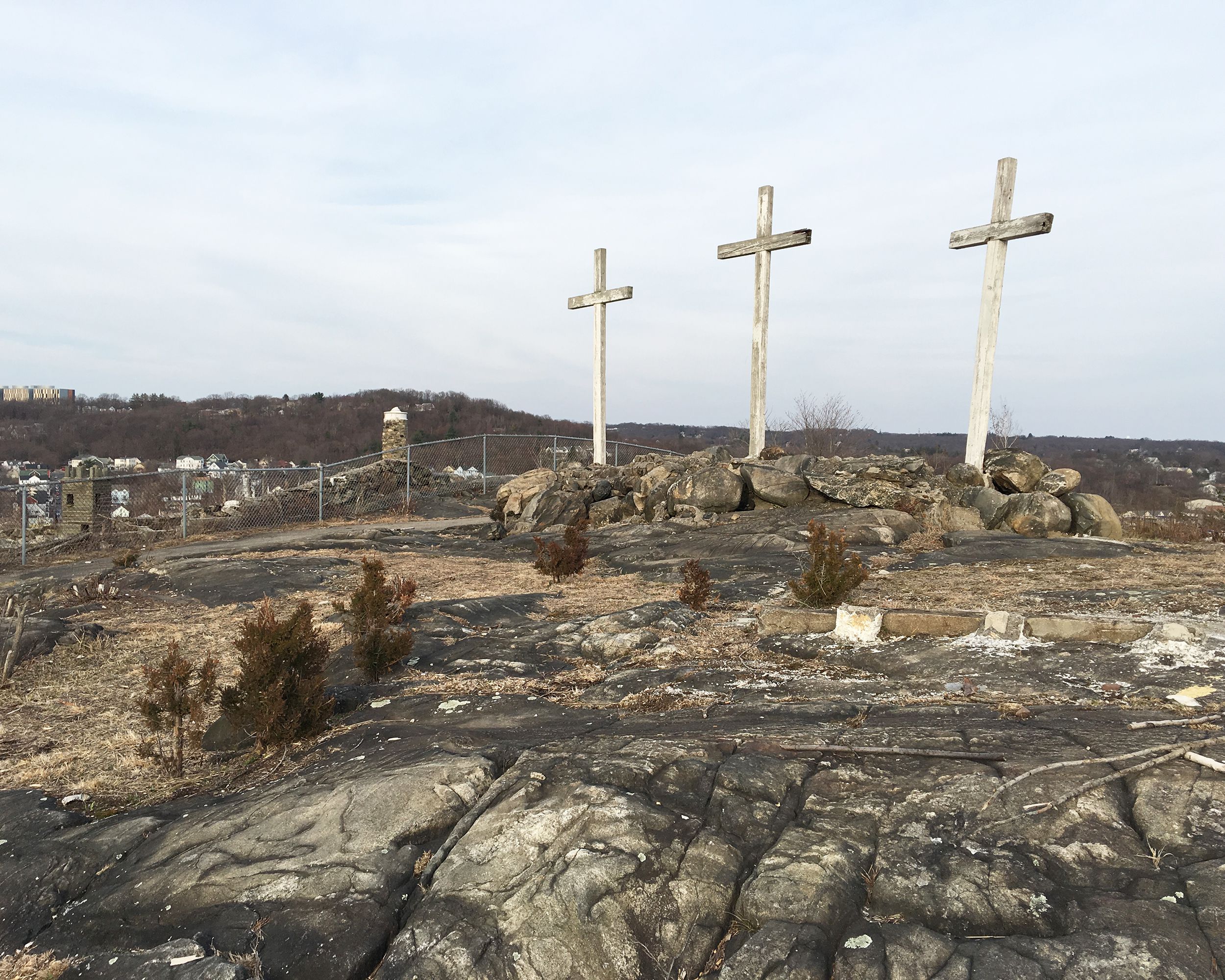 Abandoned Holy Land USA, Waterbury, Connecticut, three large white wooden crosses on rocks, rocky terrain in the foreground, town, trees on hills, and a clouds in a blue sky in the background, during winter
