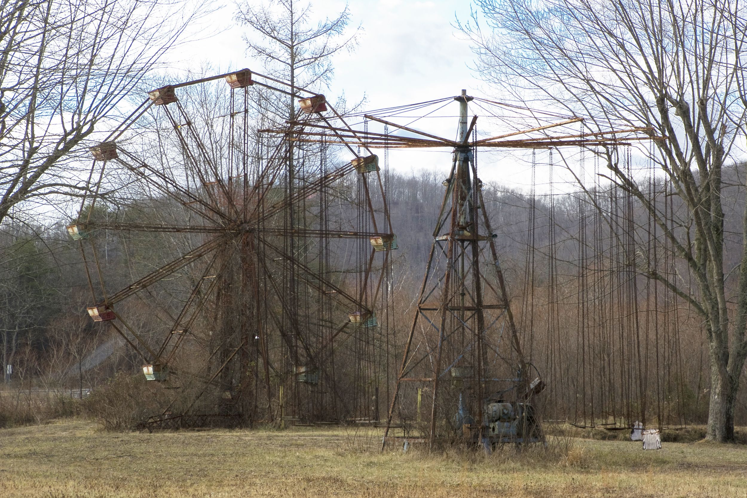 Abandoned giant wheel ride and a spinning swings ride, Lake Shawnee Amusement Park, Princeton, West Virginia, bare trees surround and in the background during winter