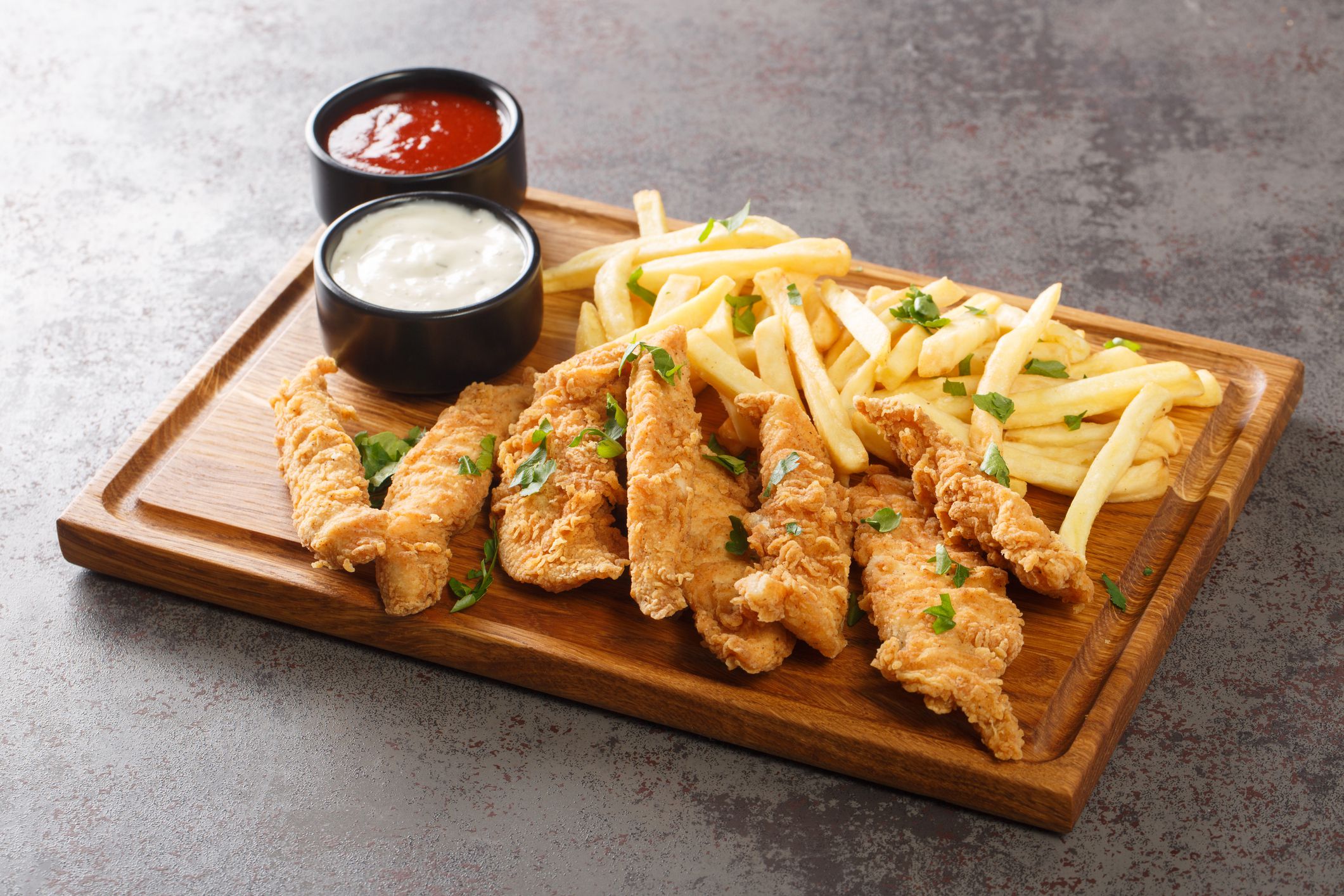 Air fried crumbed chicken tenderloins on wooden cutting board with fries and sauce