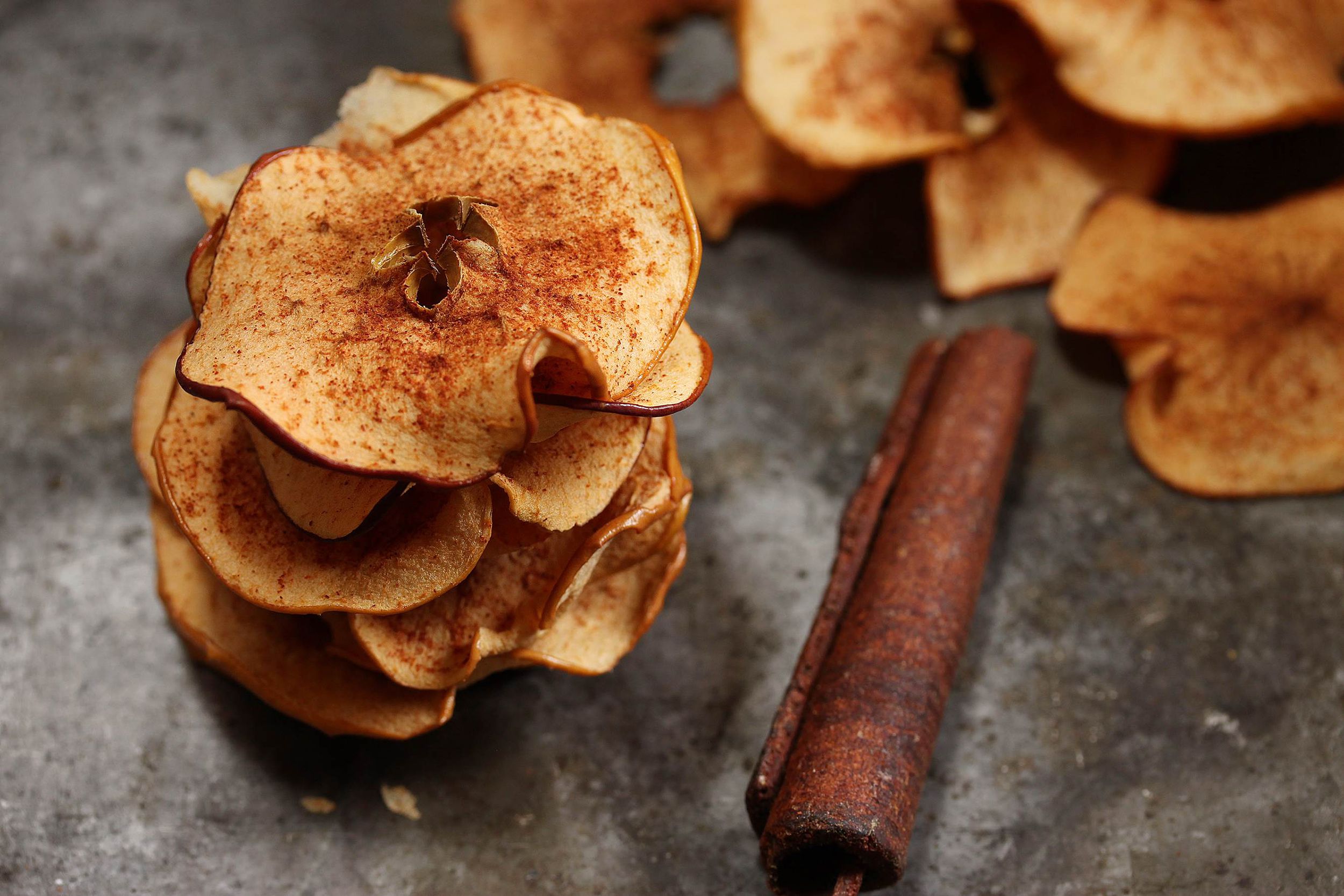 Closeup of air fryer apple chips with a cinnamon stick on grey table and apple chips in the background