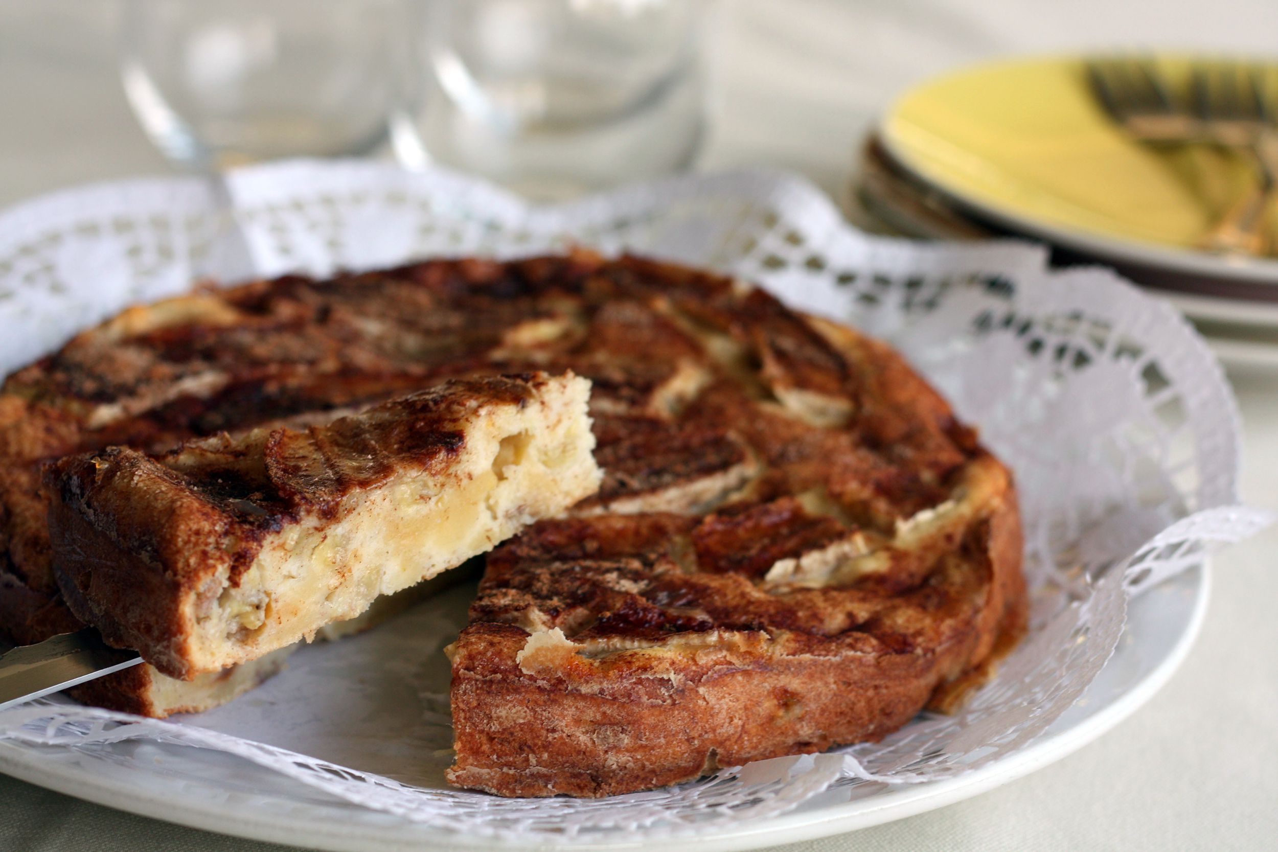 Pie server holding a slice of air fryer banana bread with rest of loaf on a white plate with plates and glasses in the background