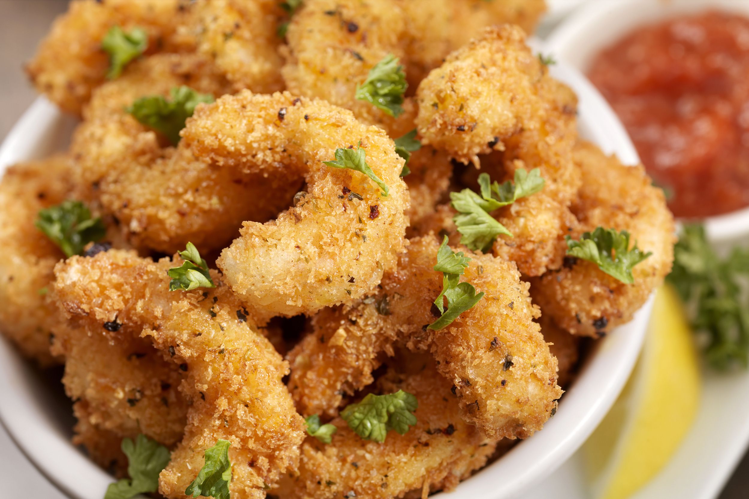 Closeup of air fryer bang bang breaded fried shrimp in a white bowl with sauce, a lemon, and herbs in the background