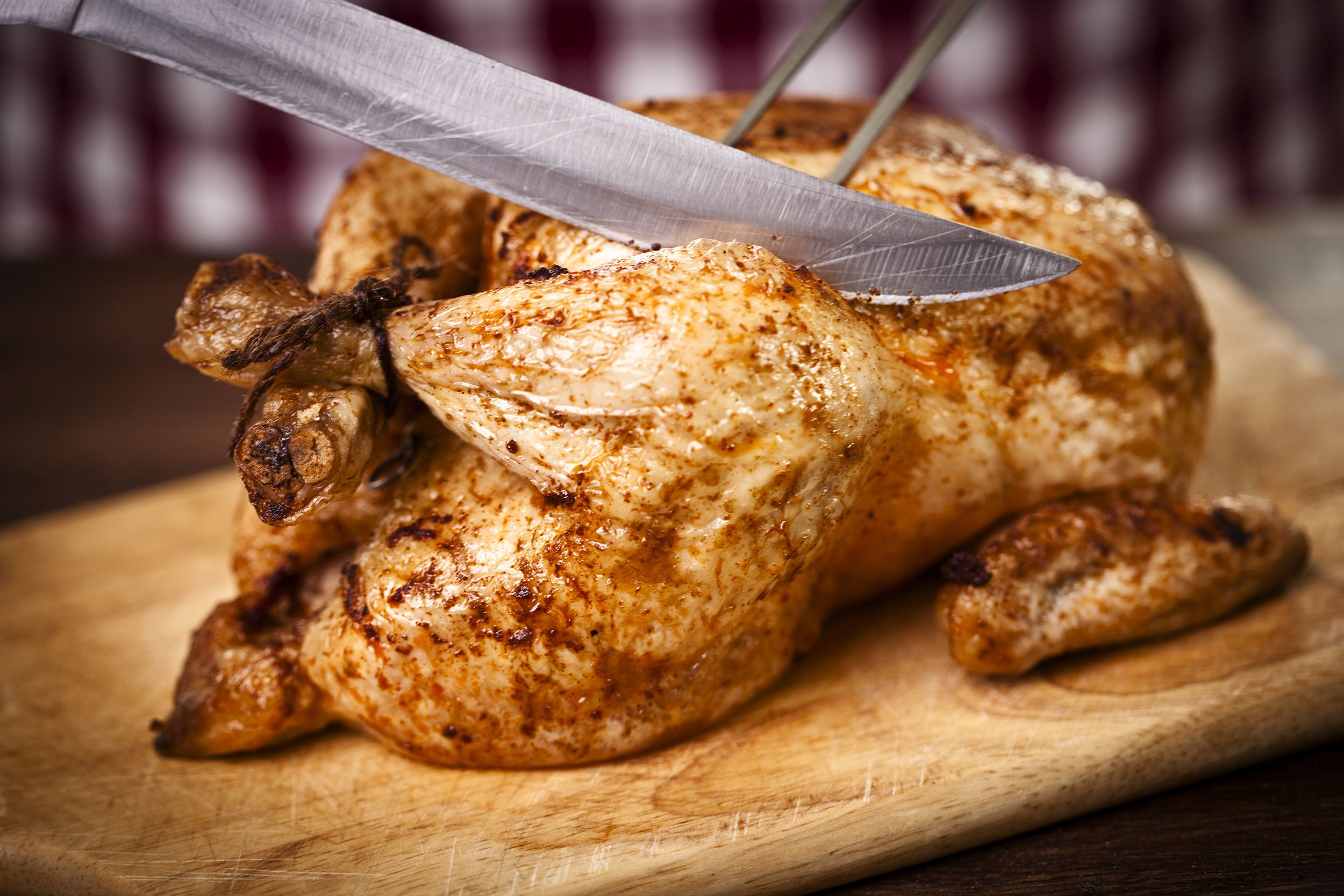 Air fryer rotisserie chicken with knife and carving fork cutting into it, on a wooden cutting board with blurred background