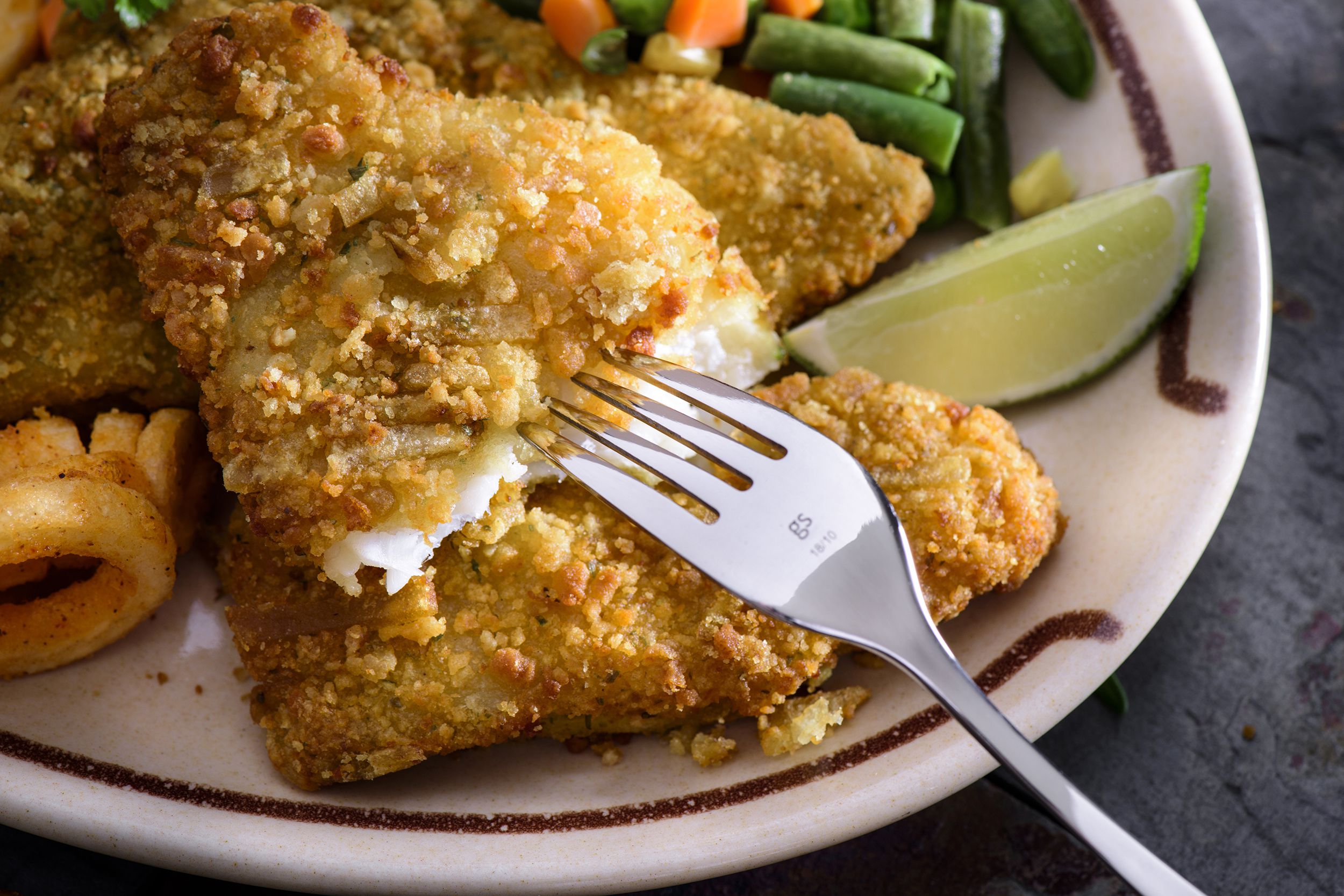 Air fryer southern style catfish with green beans with fork into a piece of catfish on a white plate with curly fries, a lime slice, and vegetables
