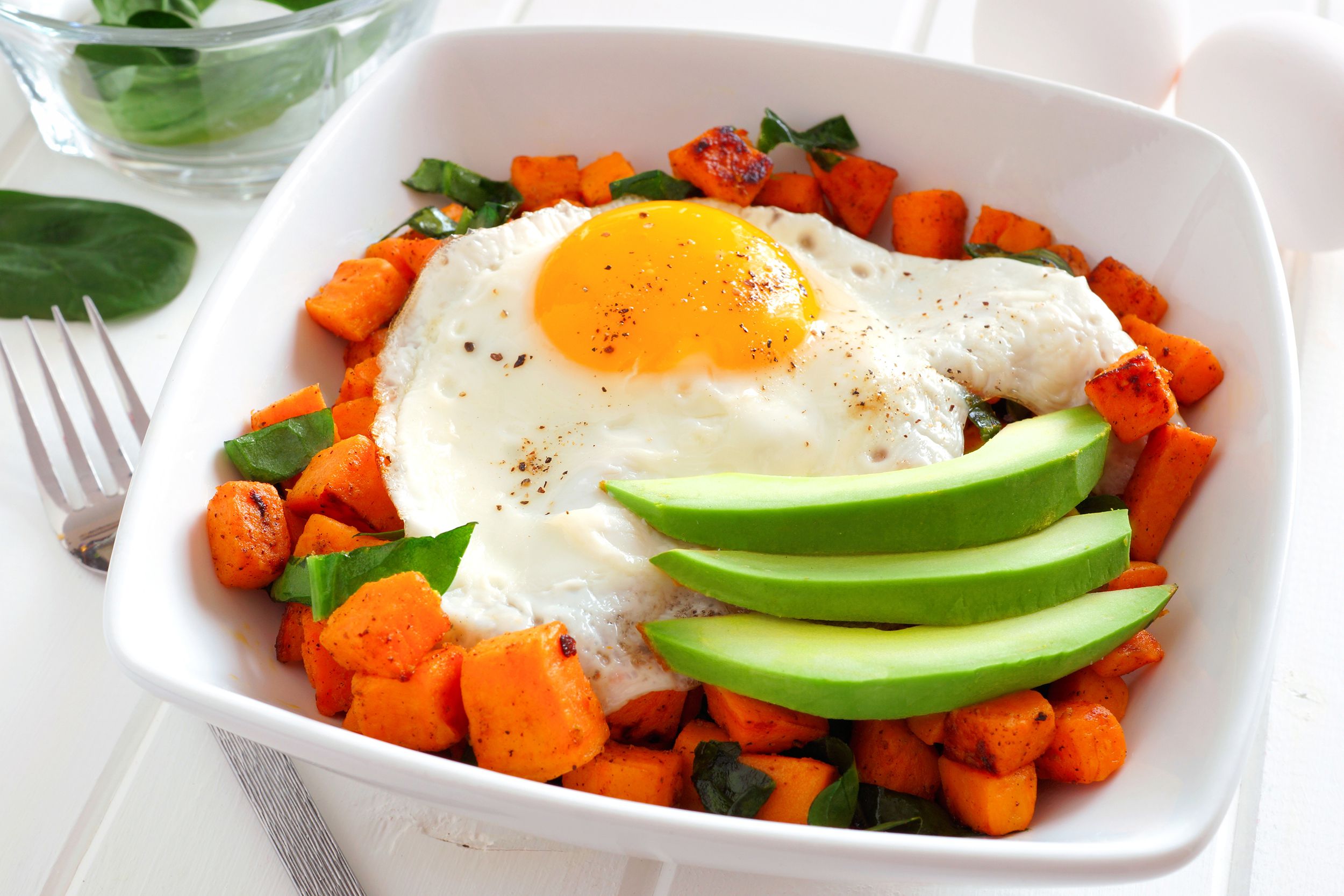 Air fryer sweet potato hash with a sunny side-up egg and slices of avocado in a white, square bowl with fork, two eggs, and herbs in the background