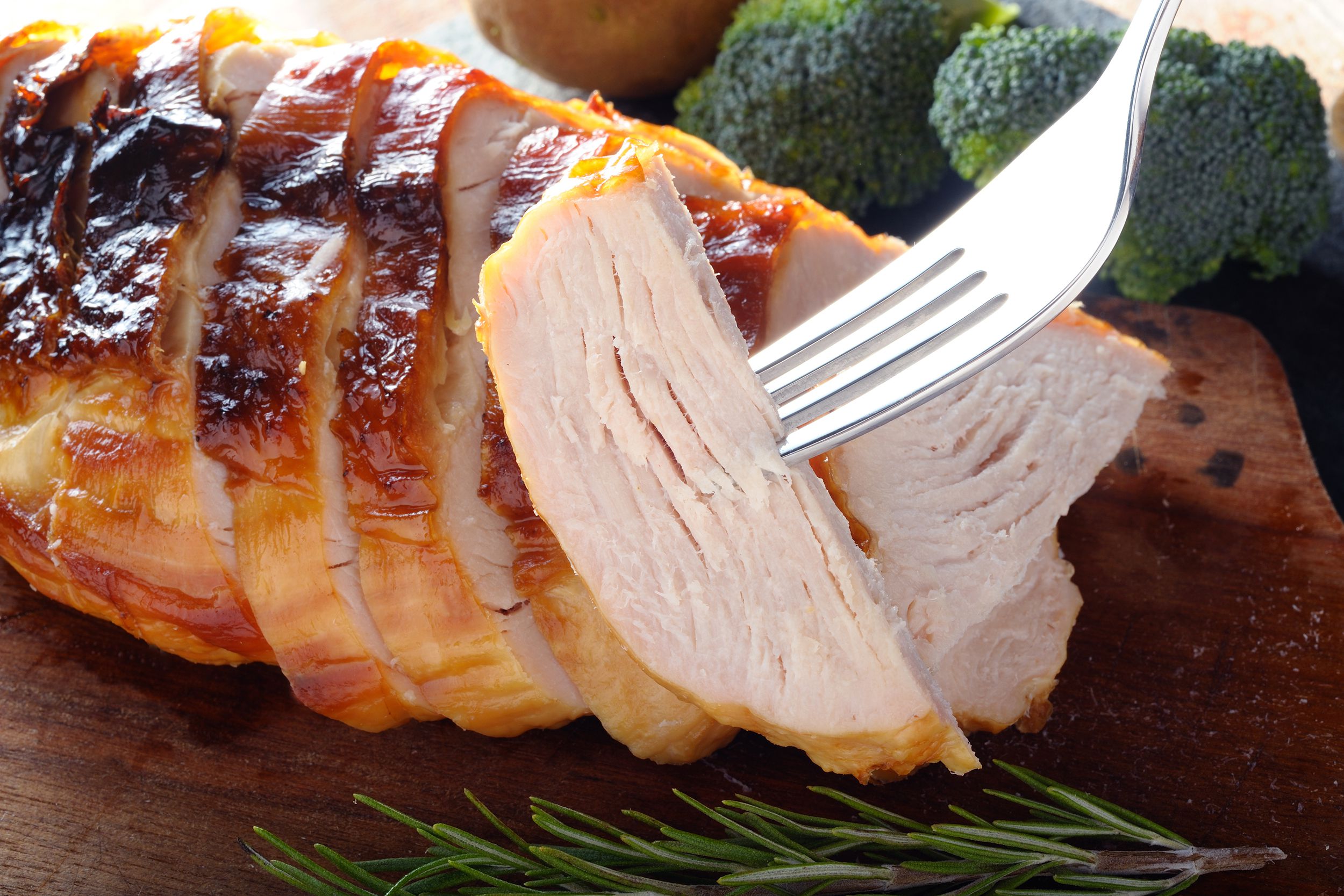Fork holding a slice of air fryer turkey breast in front of several slices with herbs, broccoli, and potato in the background