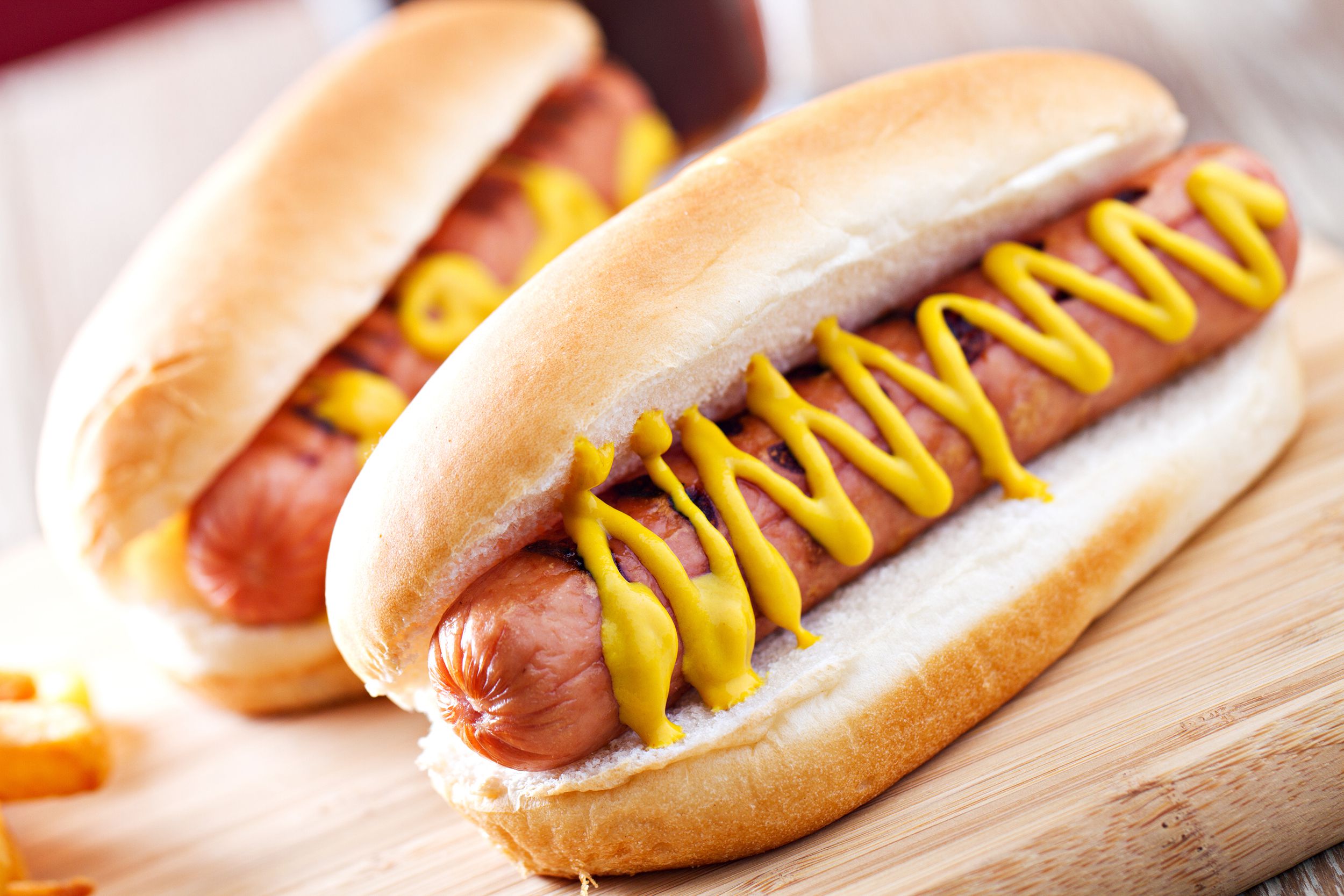 Two basic air fryer hot dogs with mustard in buns on a wooden cutting board with blurred background