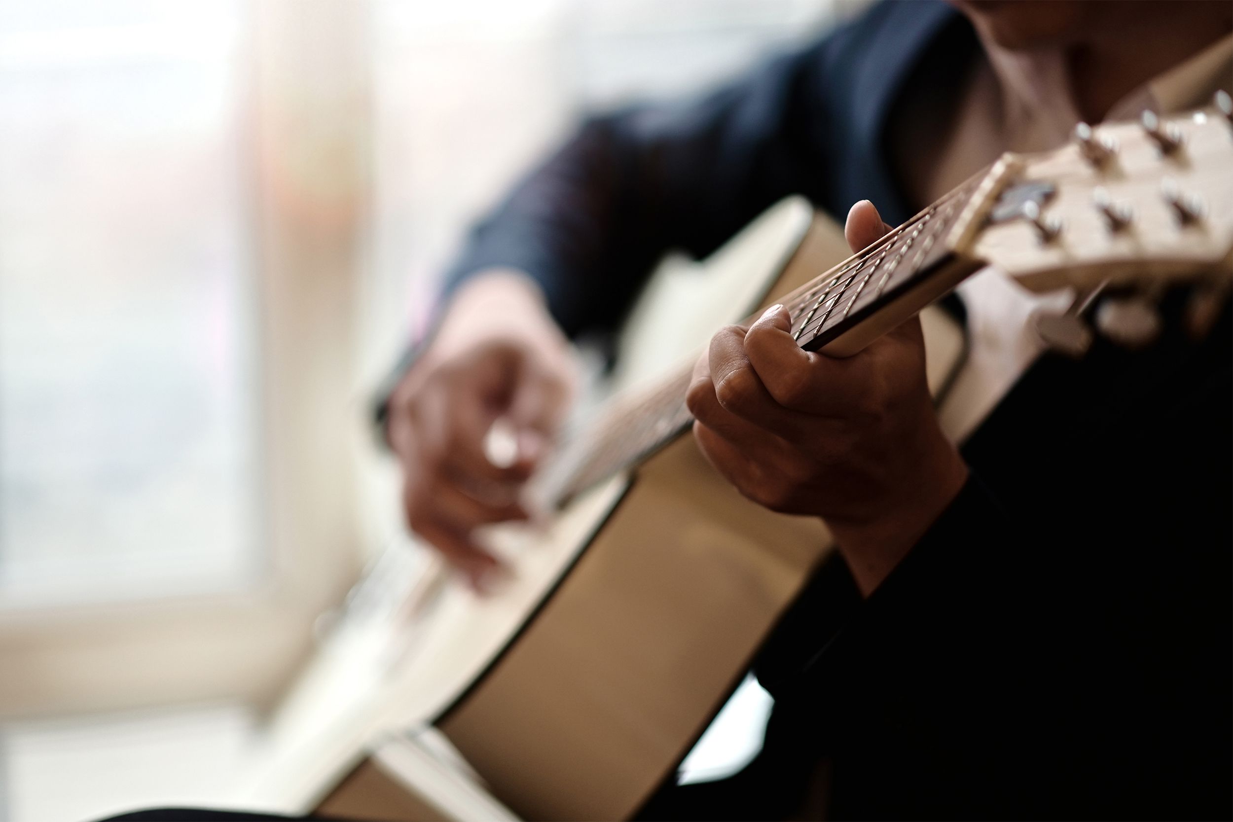 Closeup of fret of acoustic guitar as senior man is playing it with midsection of man against body of acoustic guitar, blurred background of light coming through the window