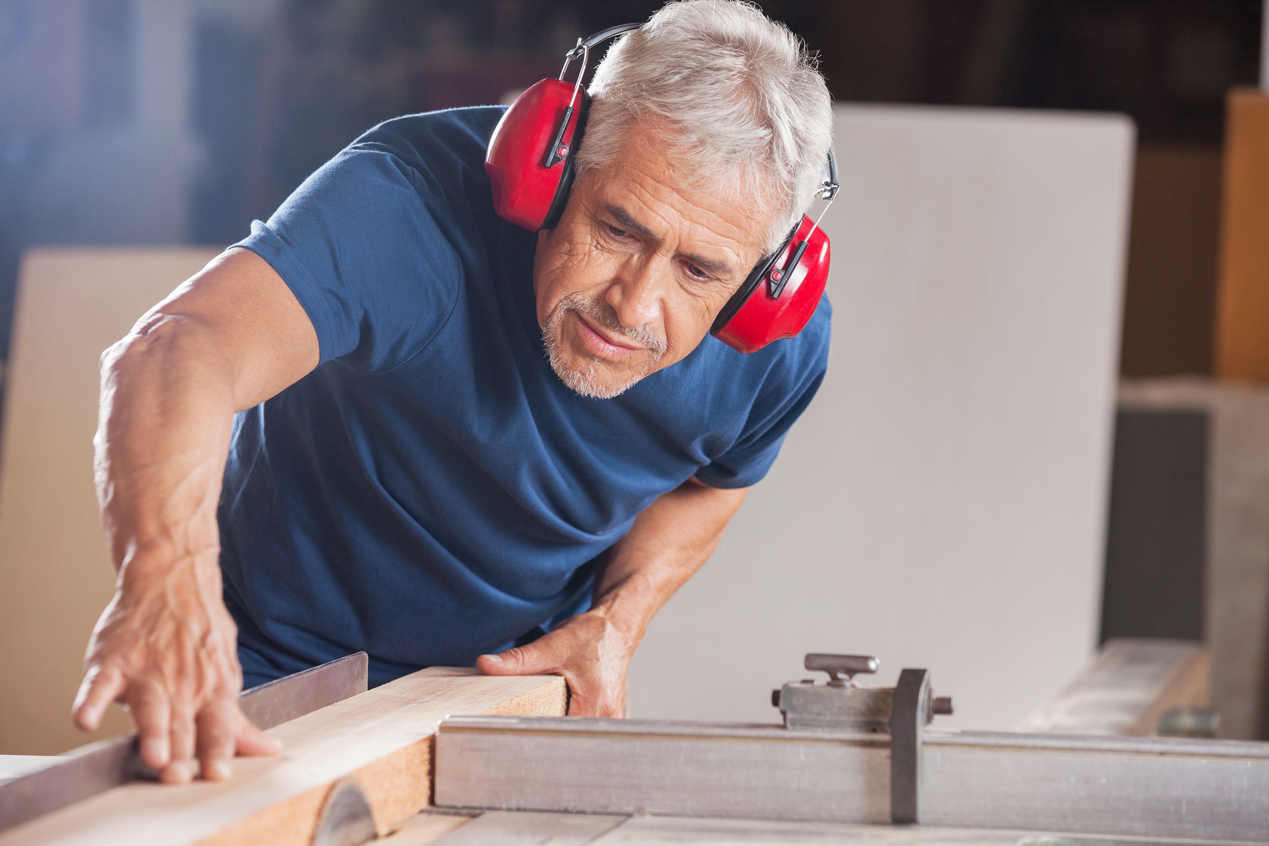 Concentrated senior male carpenter cutting a slab of wood in a workshop, with a blurred background of two large pieces of wood
