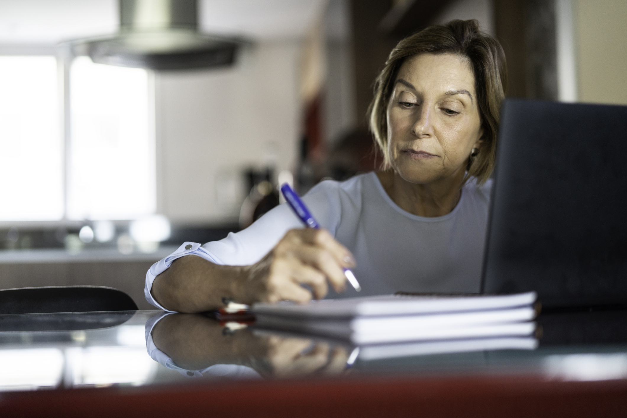 Focus on concentrated senior woman sitting at desk with laptop, writing with a pen on a notebook in living room, with blurred background of kitchen
