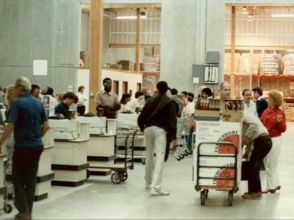 Shoppers stand in line at warehouse-style checkout counters, pushing carts filled with large items and bulk goods in a spacious warehouse store with tall concrete walls and visible storage racks.