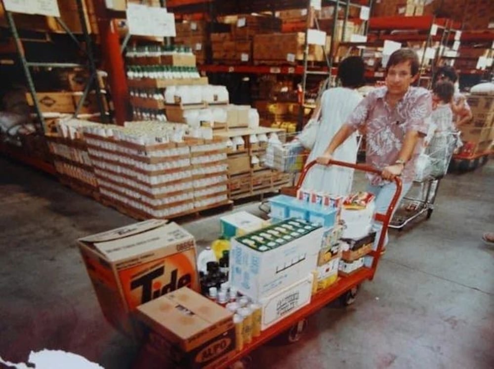 A person pushes a red flat cart loaded with bulk household items, including a large box of Tide detergent, in a warehouse store aisle with shelves stacked high with various products. Other shoppers are visible in the background.
