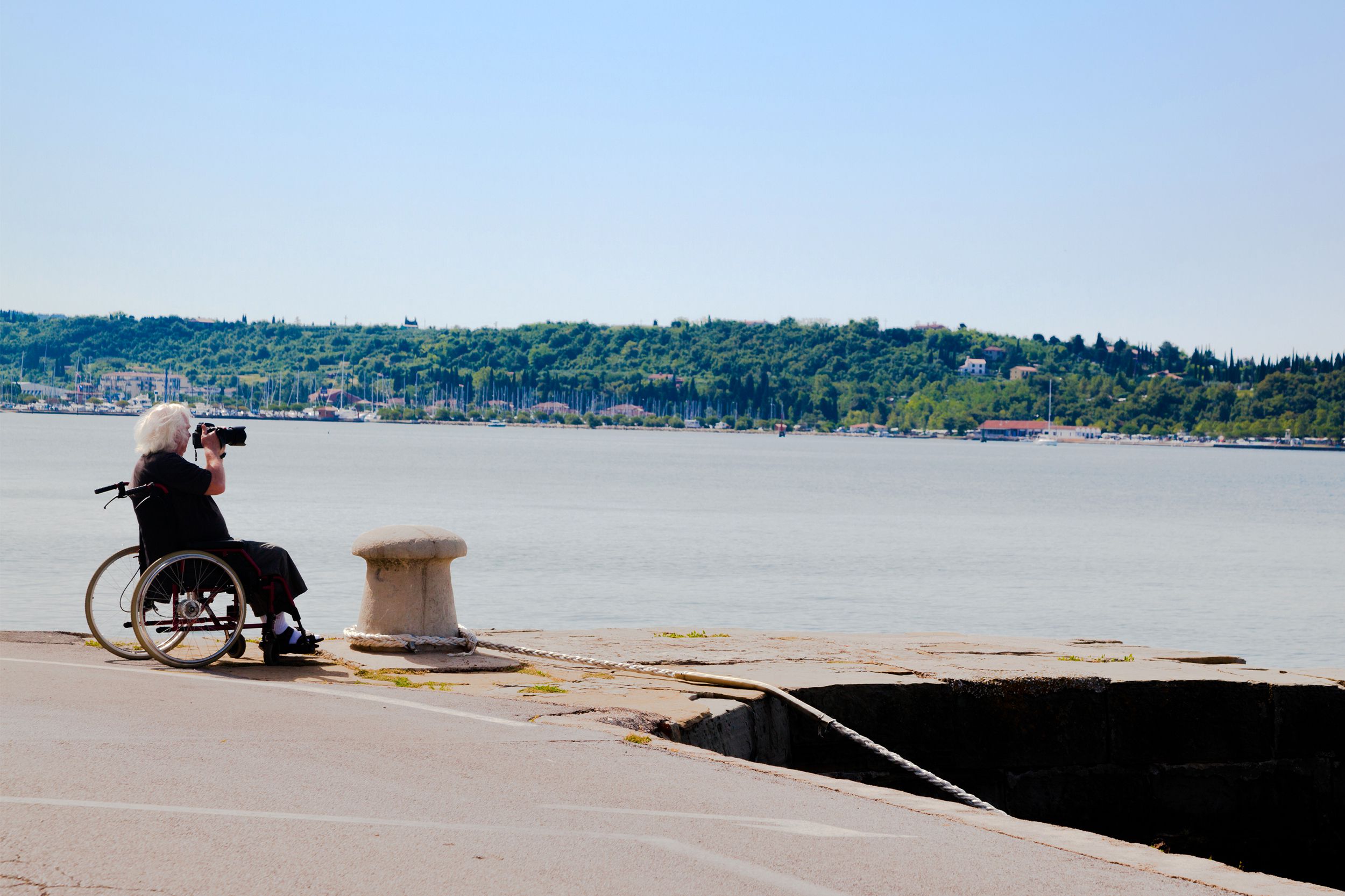 Disabled senior man in wheelchair taking photographs with dslr camera along walkway of a waterway on holiday with forest and blue sky in the background