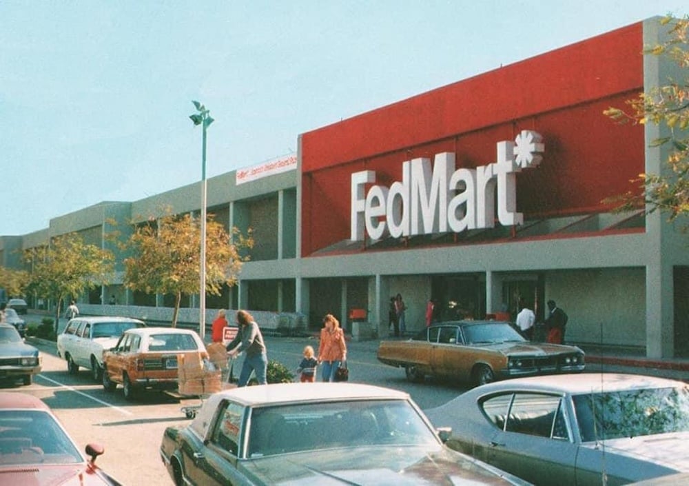 A busy parking lot outside a large FedMart store, with people walking near vintage cars and shopping carts, and a bold red and white FedMart sign on the building, likely from the 1970s.