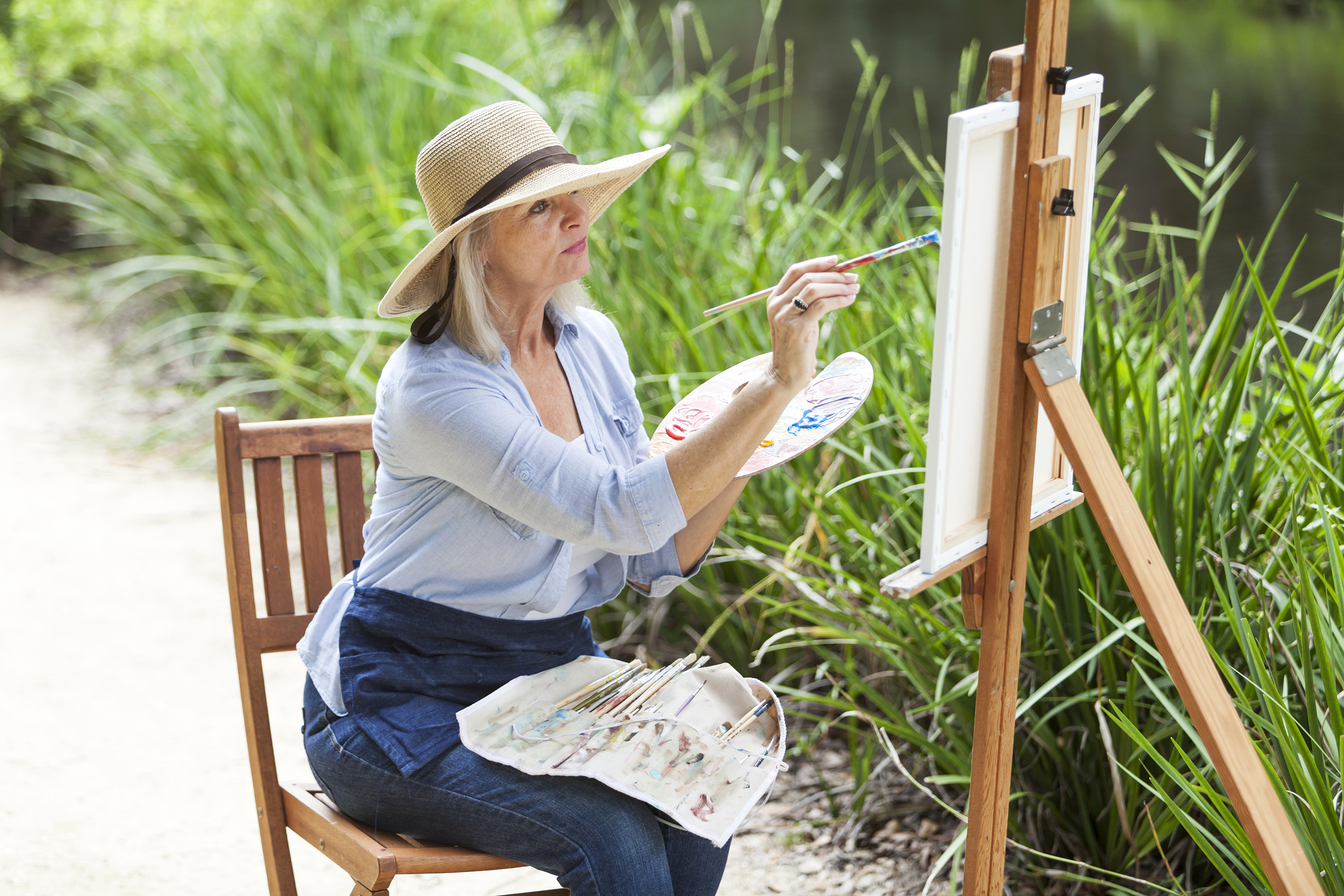 Female senior painter sitting while painting a canvas held by an easel outside doing plein air style, with a row of grass along a creek blurred in the background on a sunny day