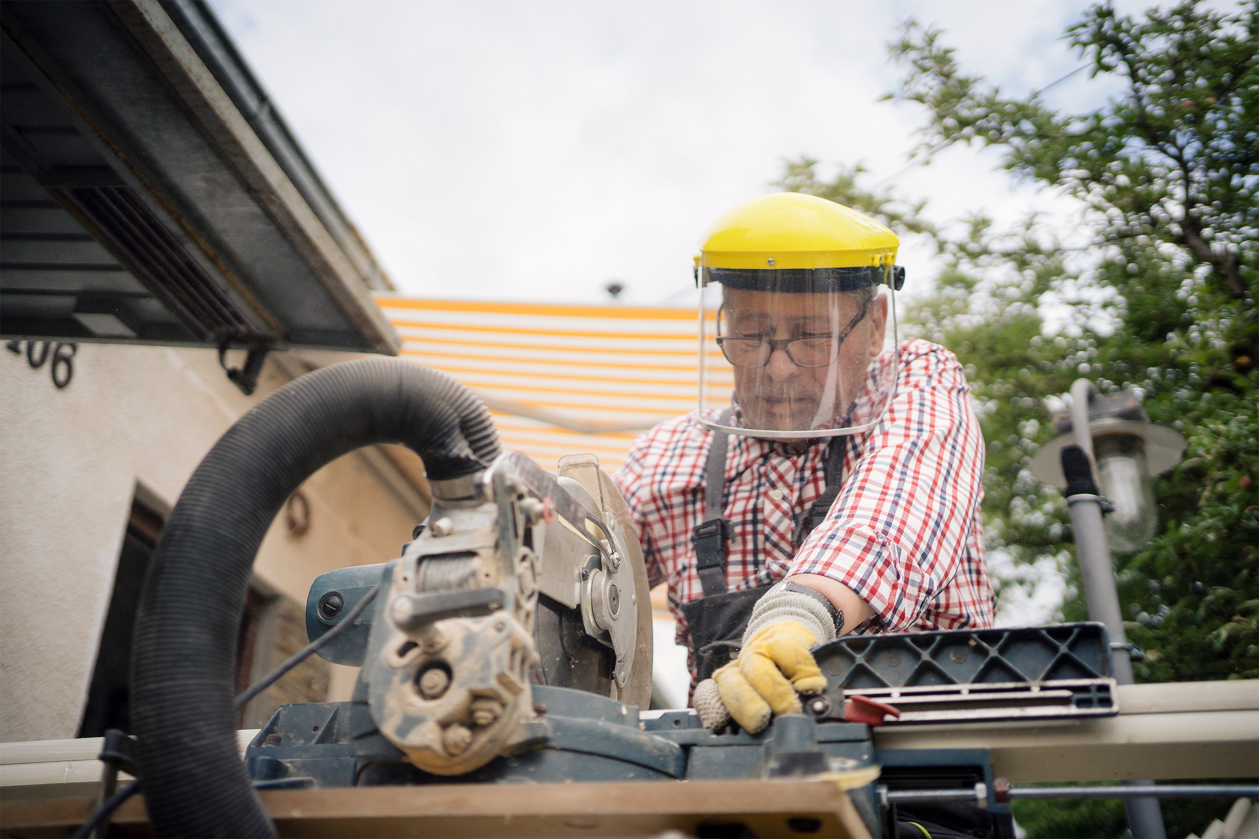 Focus on a senior man wearing protective gear while using a table saw while sawing a board of wood to use in building a home with home under construction and trees in the background