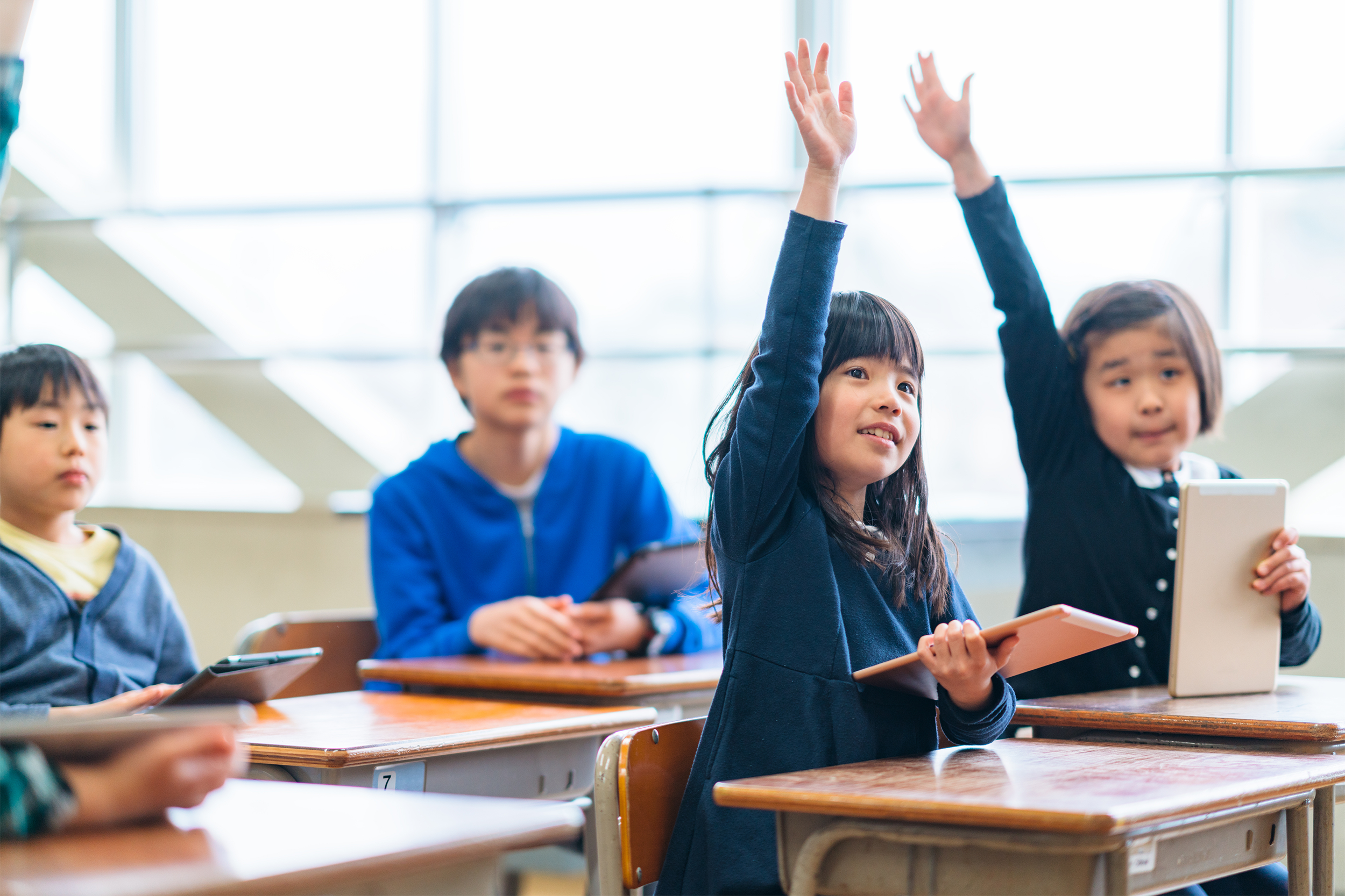 Focus on five Attentive elementary school students sitting at desks, engaged in a lecture in a classroom, three have raised hands, with a blurred background of windows