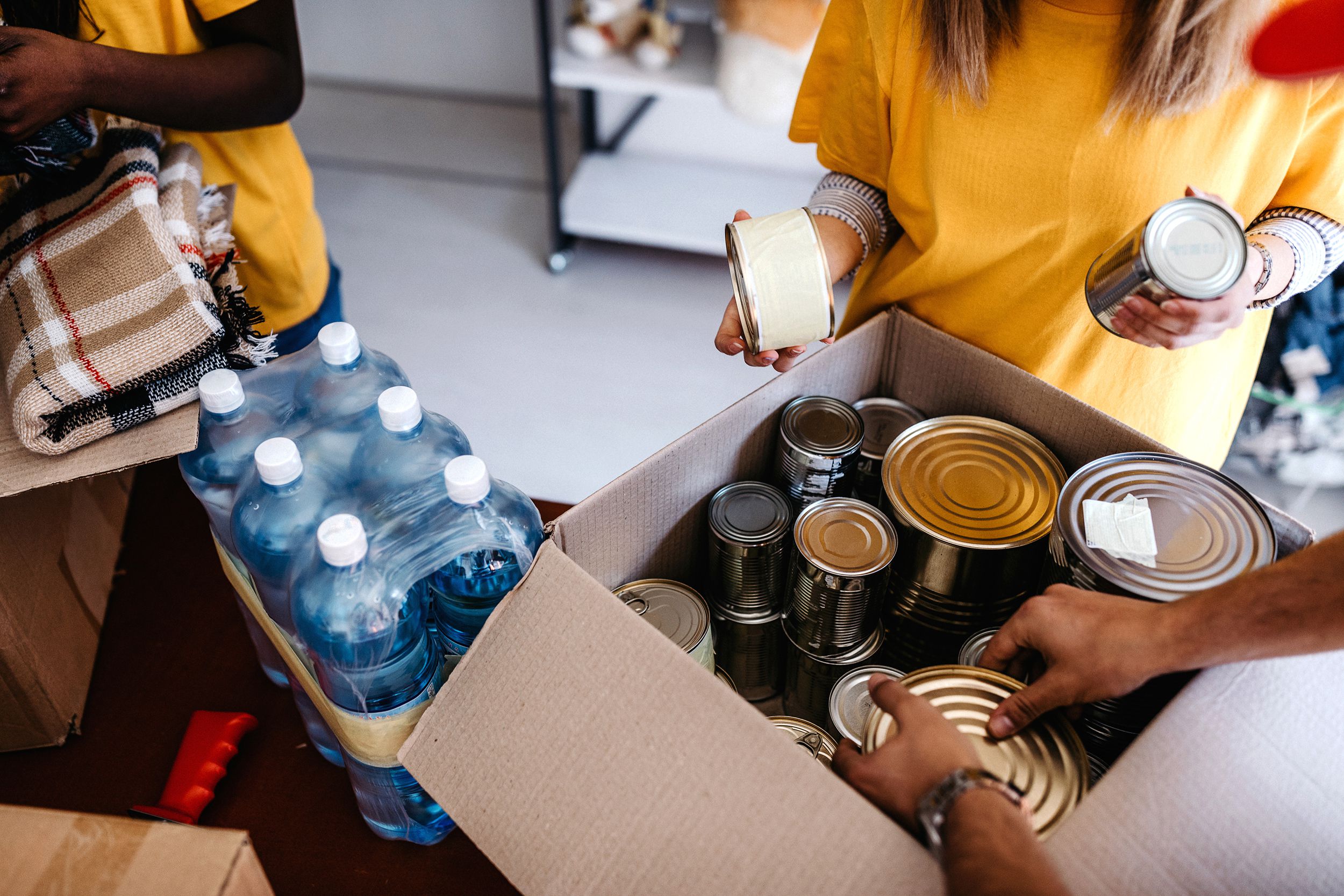Focus on hands of volunteers putting cans into boxes, one is folding a blanket, large bottles of water and other supplies on floor
