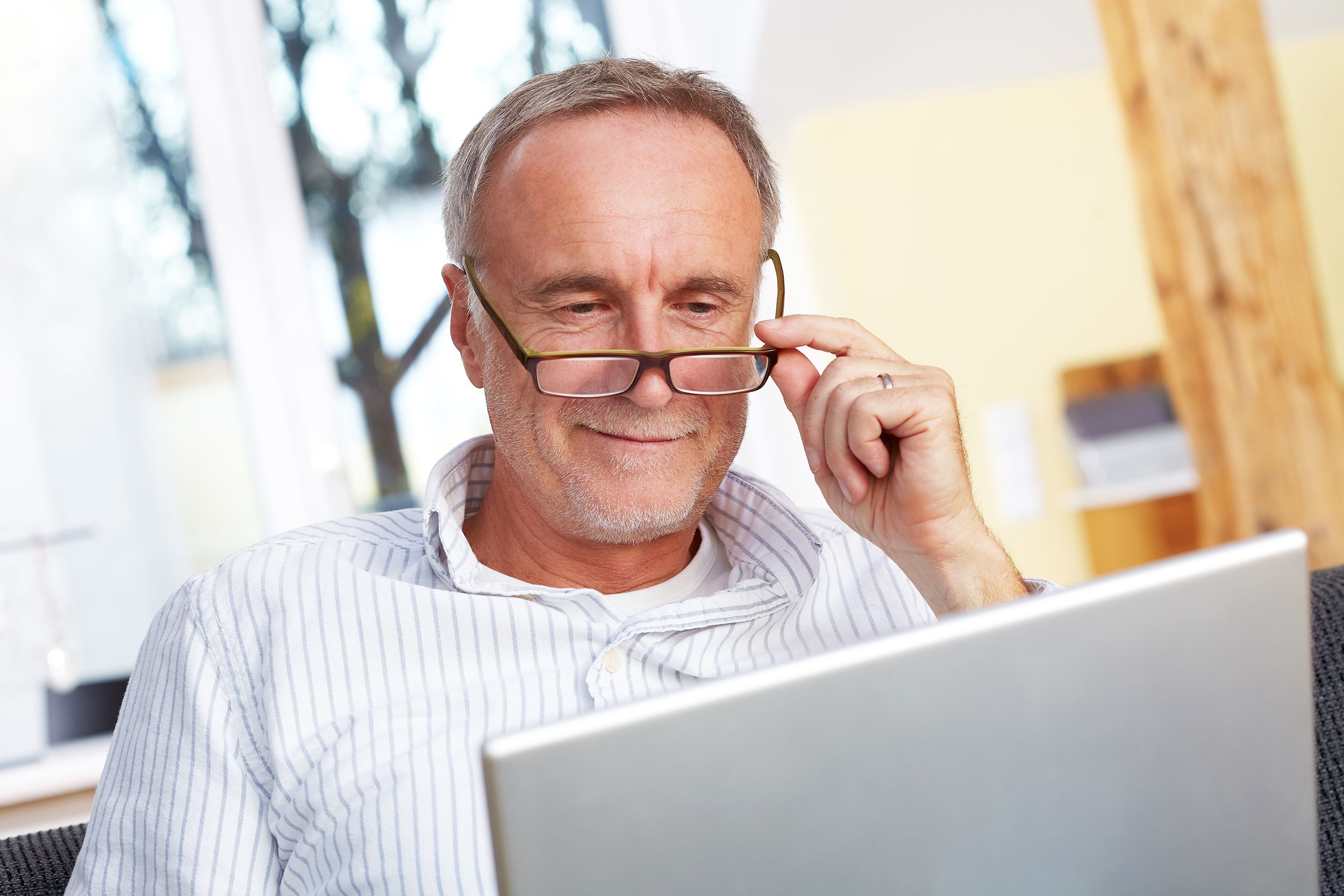 Focus on senior man touching his eyeglasses while looking at a laptop, back of laptop facing camera, with a blurred background of the room of a library