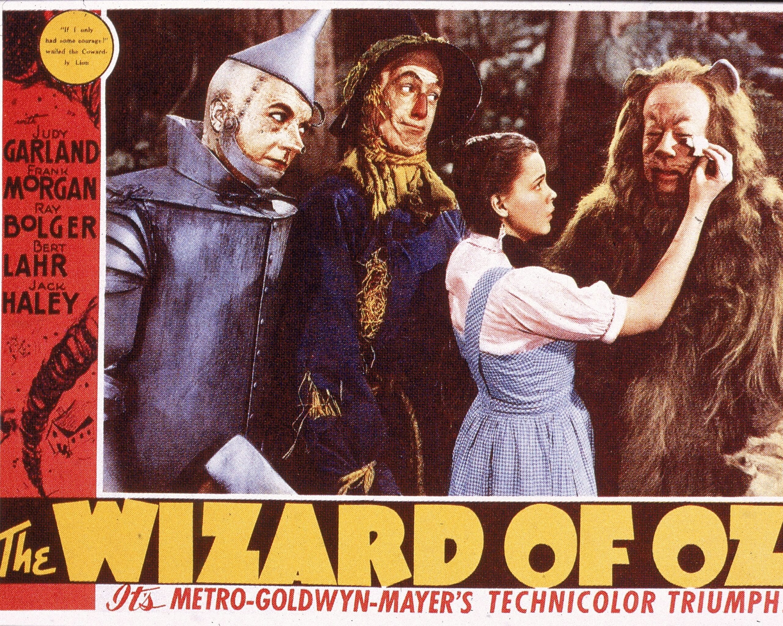 A lobby card from the film 'The Wizard Of Oz,' shows a film still of a scene in which American actress Judy Garland (1922 - 1969) (as Dorothy) wipes tears from the eyes of actor Bert Lahr (1895 - 1967) (as the Cowardly Lion), while watched by Jack Haley (