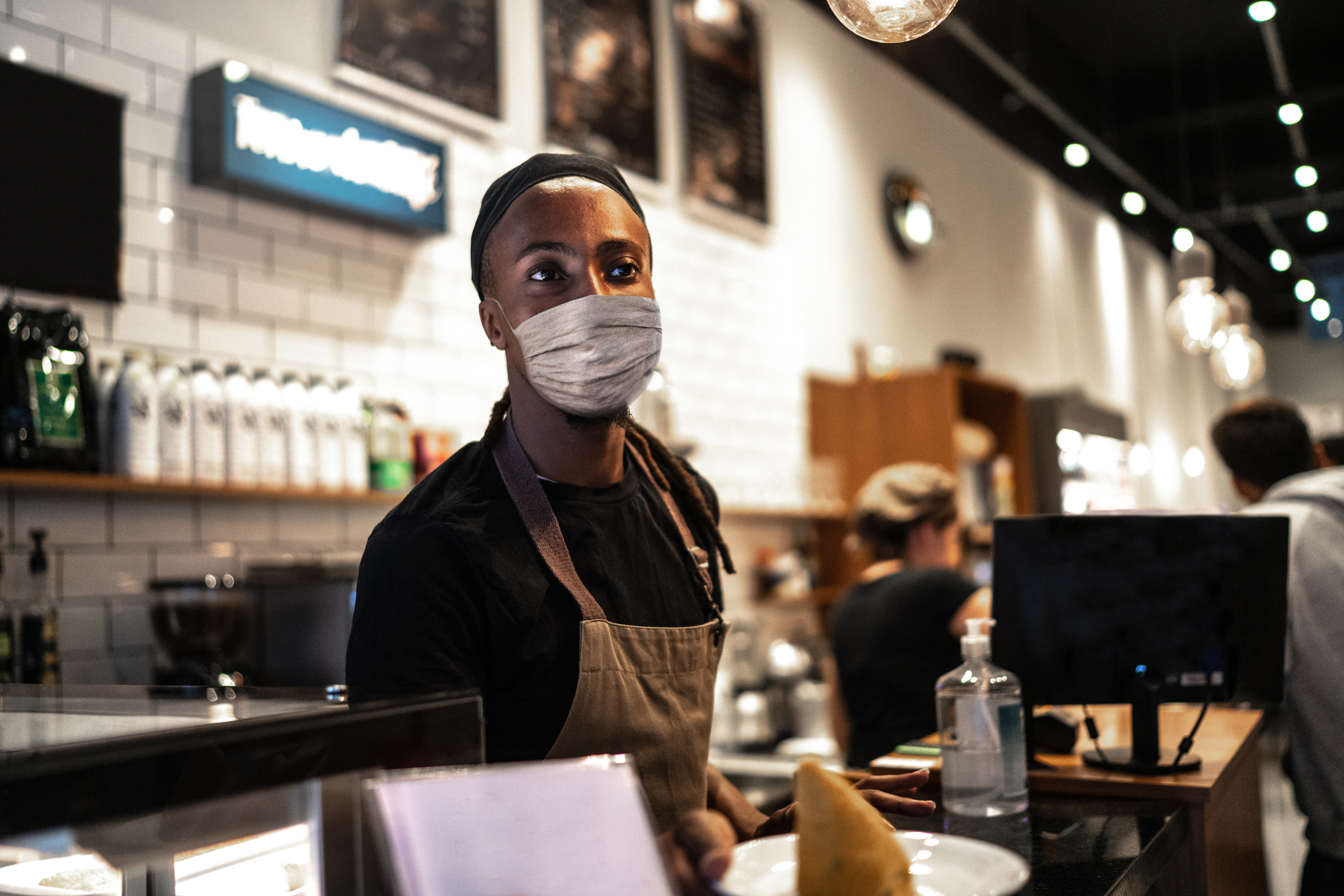 Male barista working behind the counter, wearing a face mask, selective focus, surrounded by coffee beans, products, and paraphernalia, blurred background of behind the counter of a coffee shop