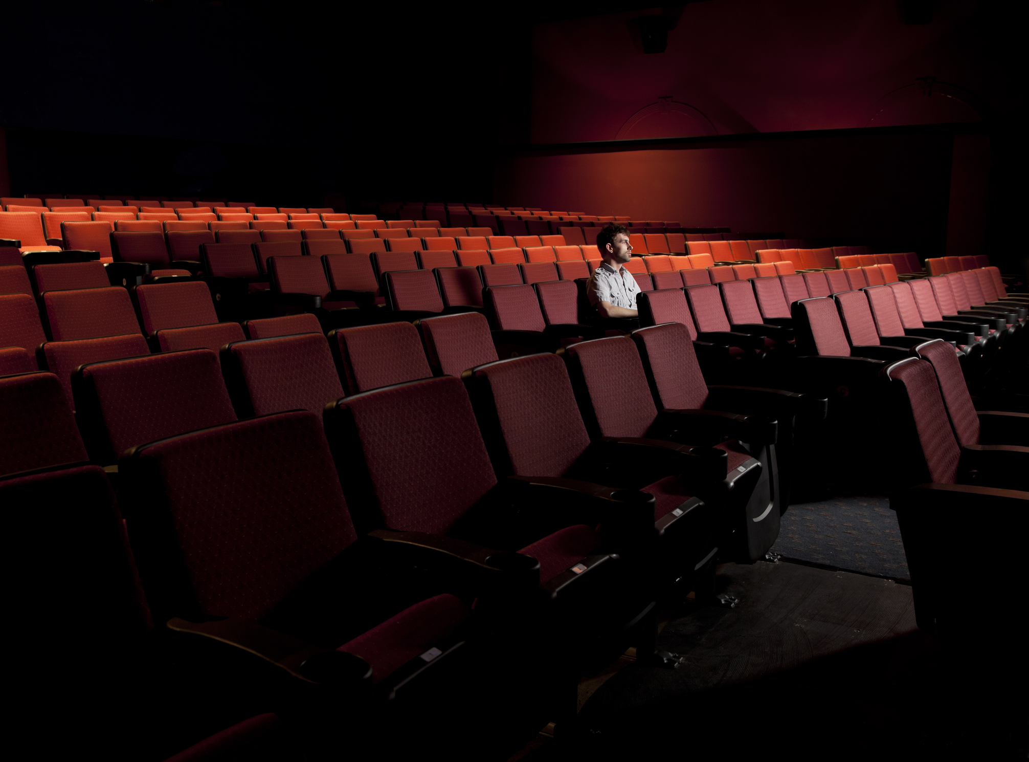Man sitting alone in a movie theater, as seen from the slight side, surrounded by empty red seats, slight light before a movie starts