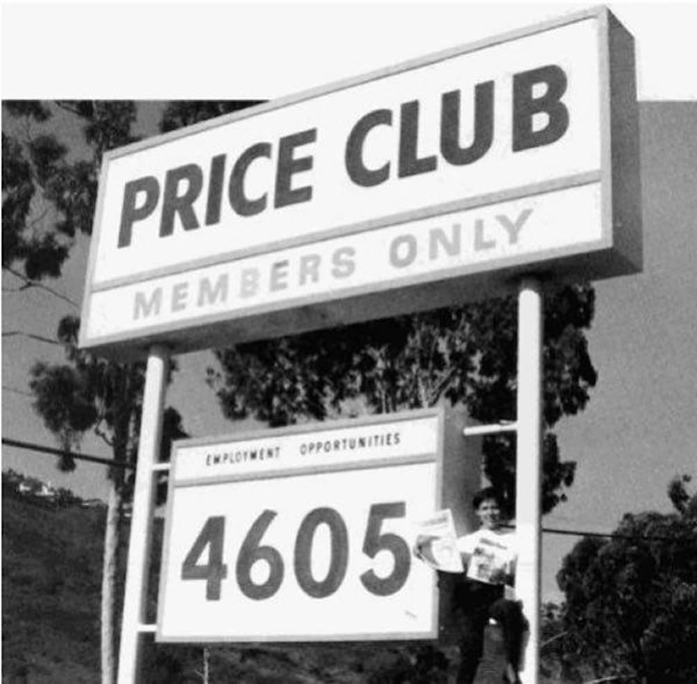 A black and white photo of a large "PRICE CLUB MEMBERS ONLY" sign with a smaller sign below reading "EMPLOYMENT OPPORTUNITIES 4605." A person stands next to the sign holding papers. Trees are visible in the background.