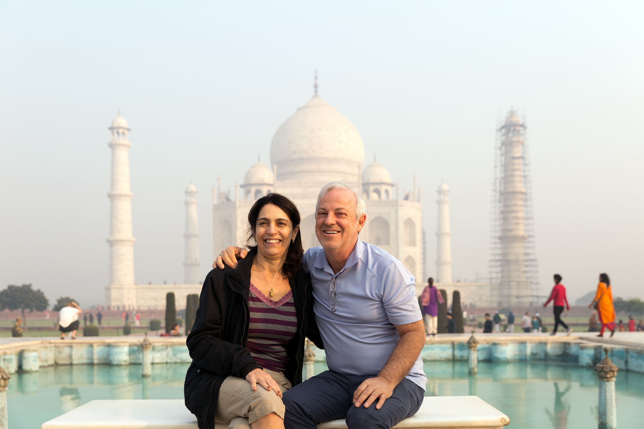 Senior couple sitting together, having photo taken in front of the reflecting pool with the Taj Mahal in the background, Agra, India on a hazy day with blue sky