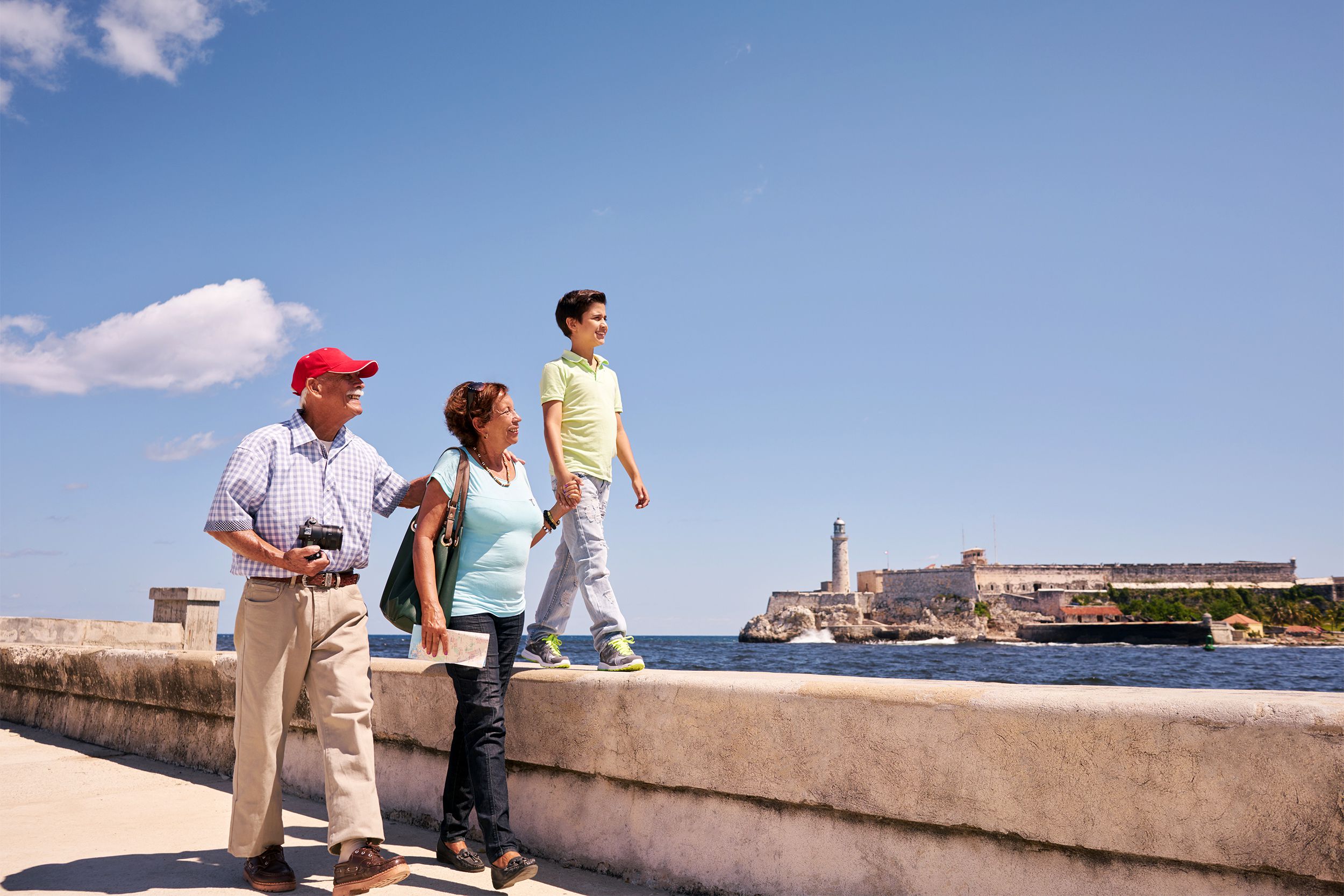 Senior couple walking with a young boy along the Malecón in Havana, Cuba on a bright sunny day with fort in the background, against a blue sky