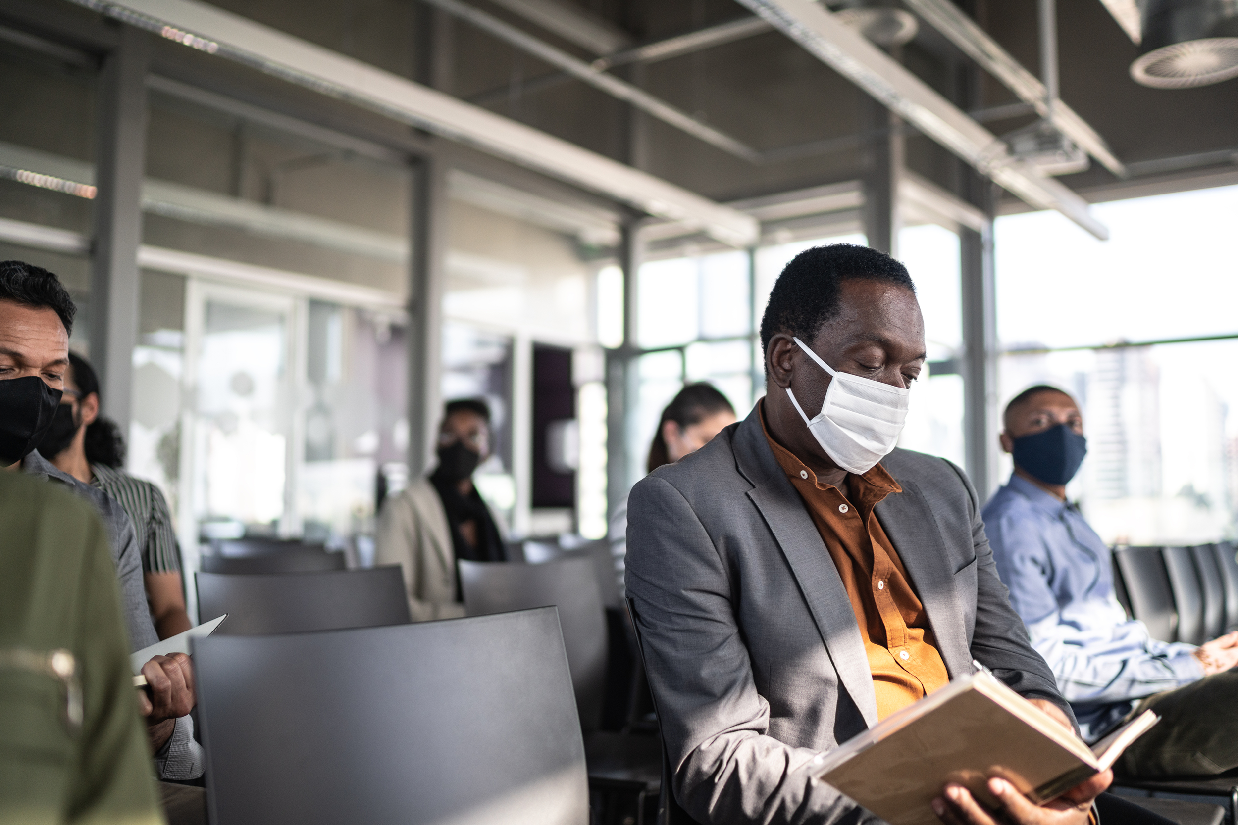 Senior man reading, sitting with other people, attending a seminar, all are wearing face masks, several empty chairs, in a modern conference room with skyscrapers in the windows, blurred background