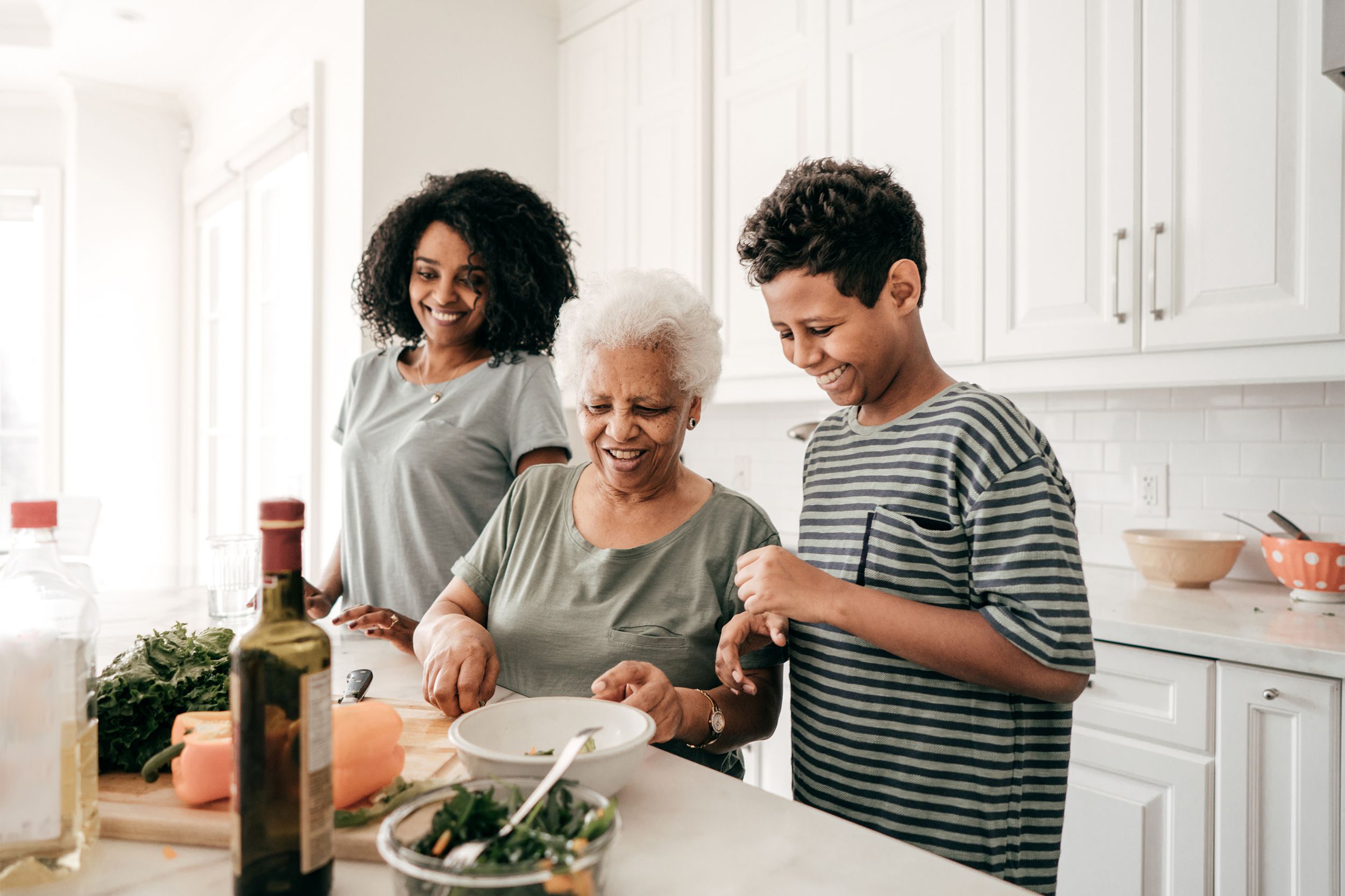 Senior woman cooking with her daughter and grandson on an island in a home kitchen, all are smiling, white kitchen background