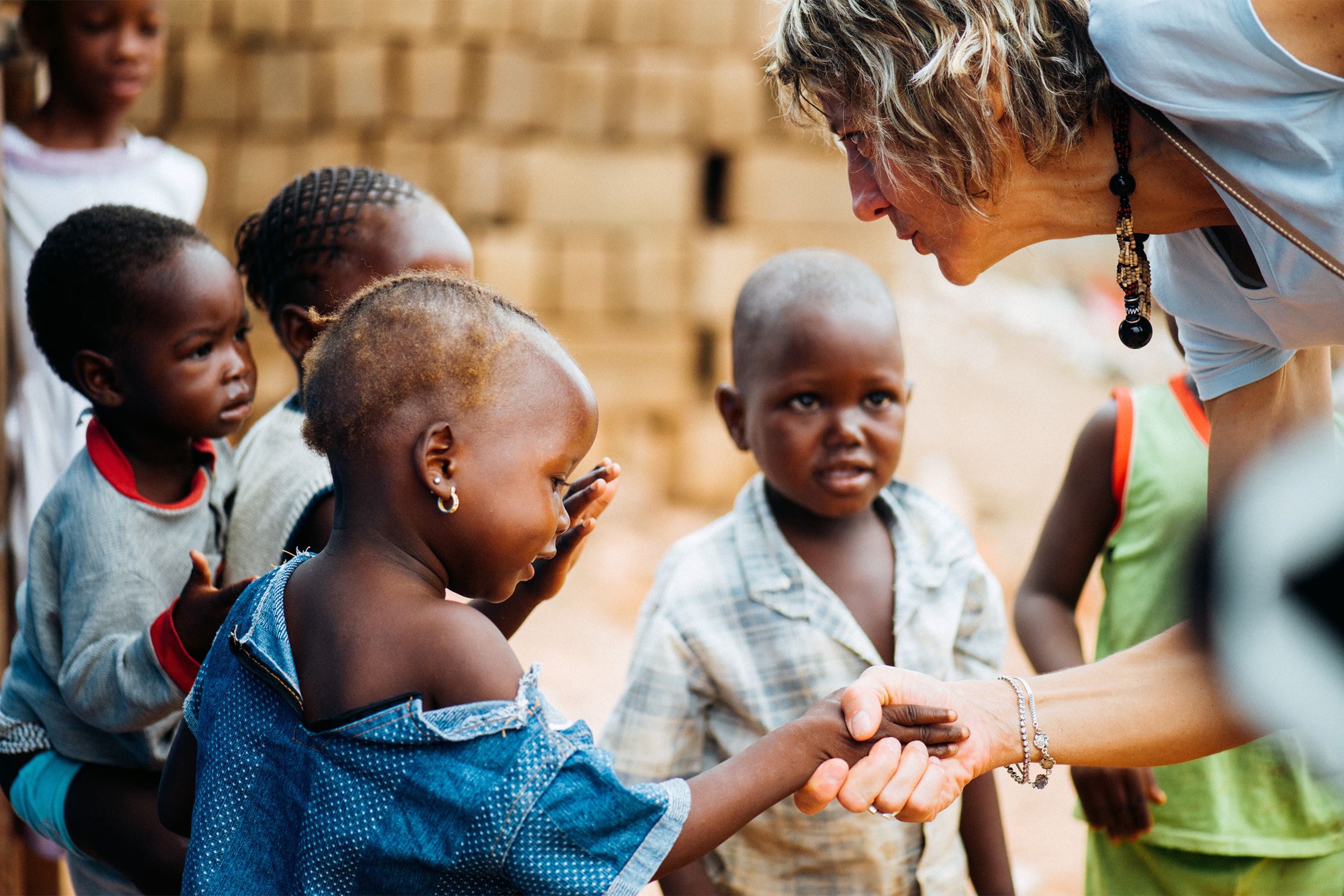 Senior woman shaking hands while talking to one poor, young African preschool boy, surrounded by a group of such, in a rural village near Bamako, Mali, with a blurred background of clay brick building