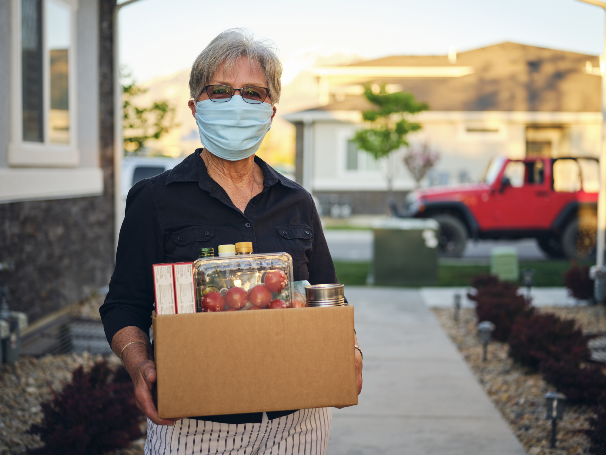 Senior woman, wearing a face mask, holding a box of delivery groceries in front of home to be delivered, street and other homes in neighborhood blurred in the background