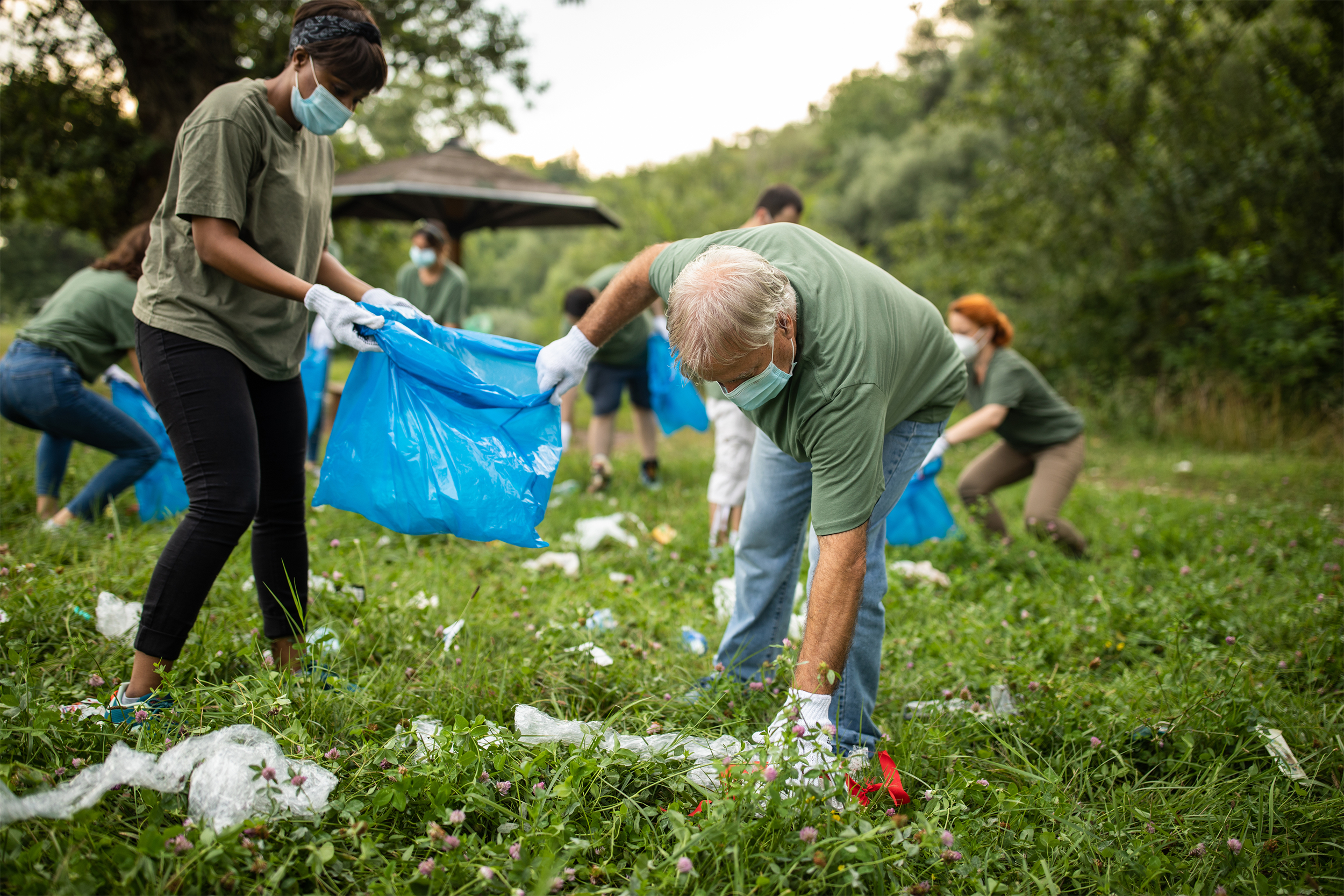 Several volunteers, wearing face masks, picking up trash in park, focus on two male volunteers, with trees and forest blurred in the background