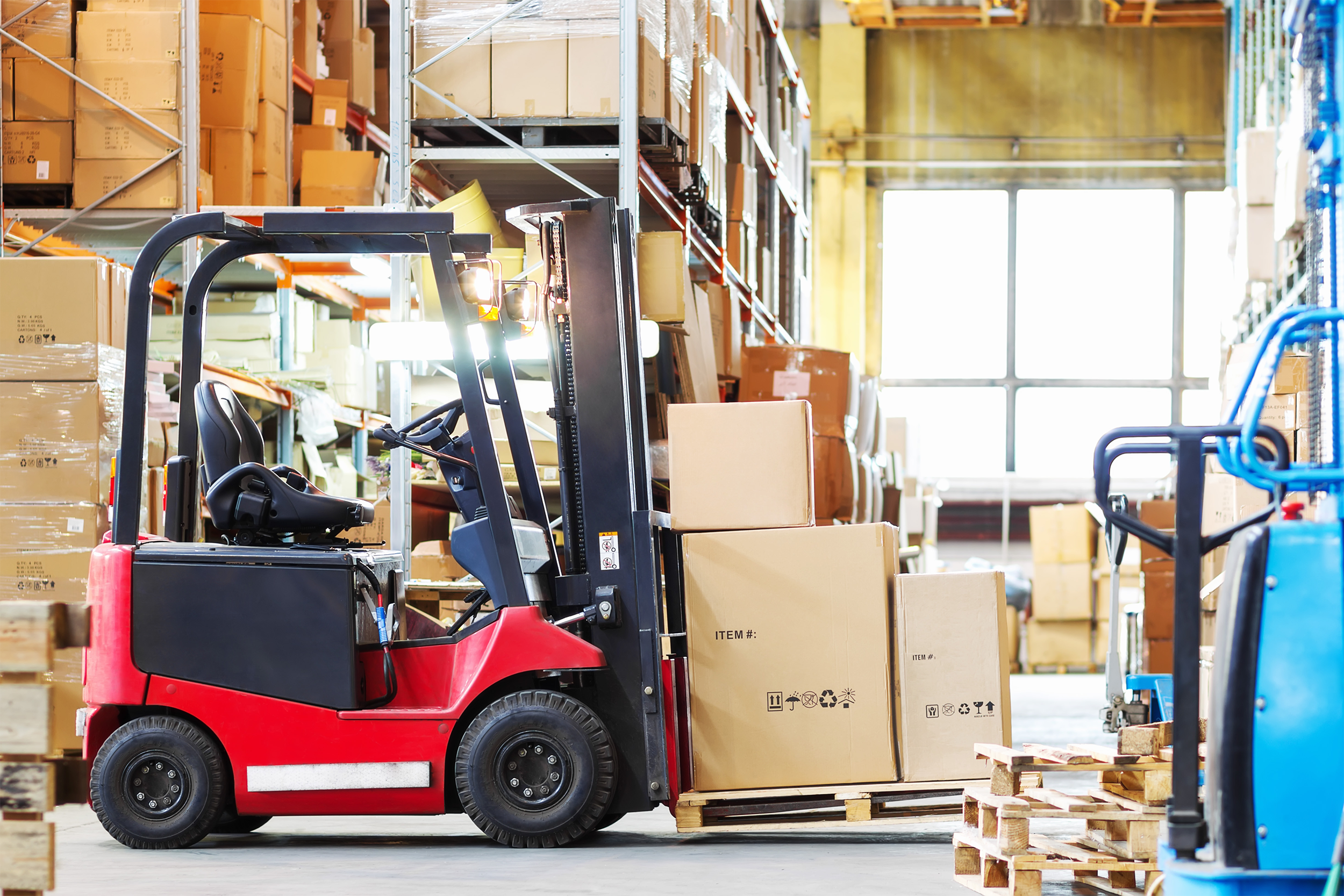 Sideview of a red warehouse forklift with three large cardboard boxes in a modern warehouse, surrounded by numerous large cardboard boxes organized in shelves