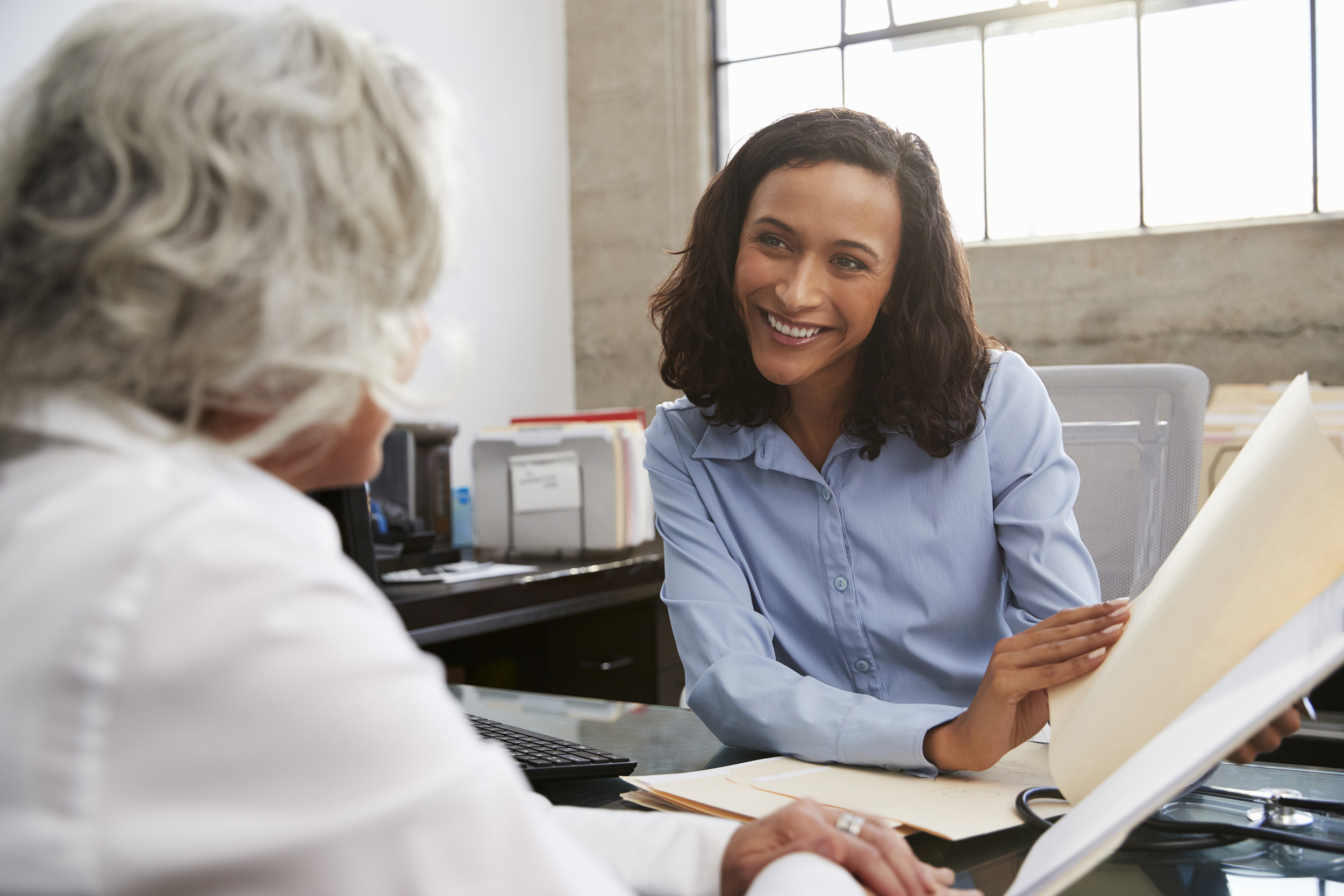 Smiling female analyst in consultation with a senior woman at her desk, back of senior and front of analyst, files and organized paperwork, wall and windows blurred in the background