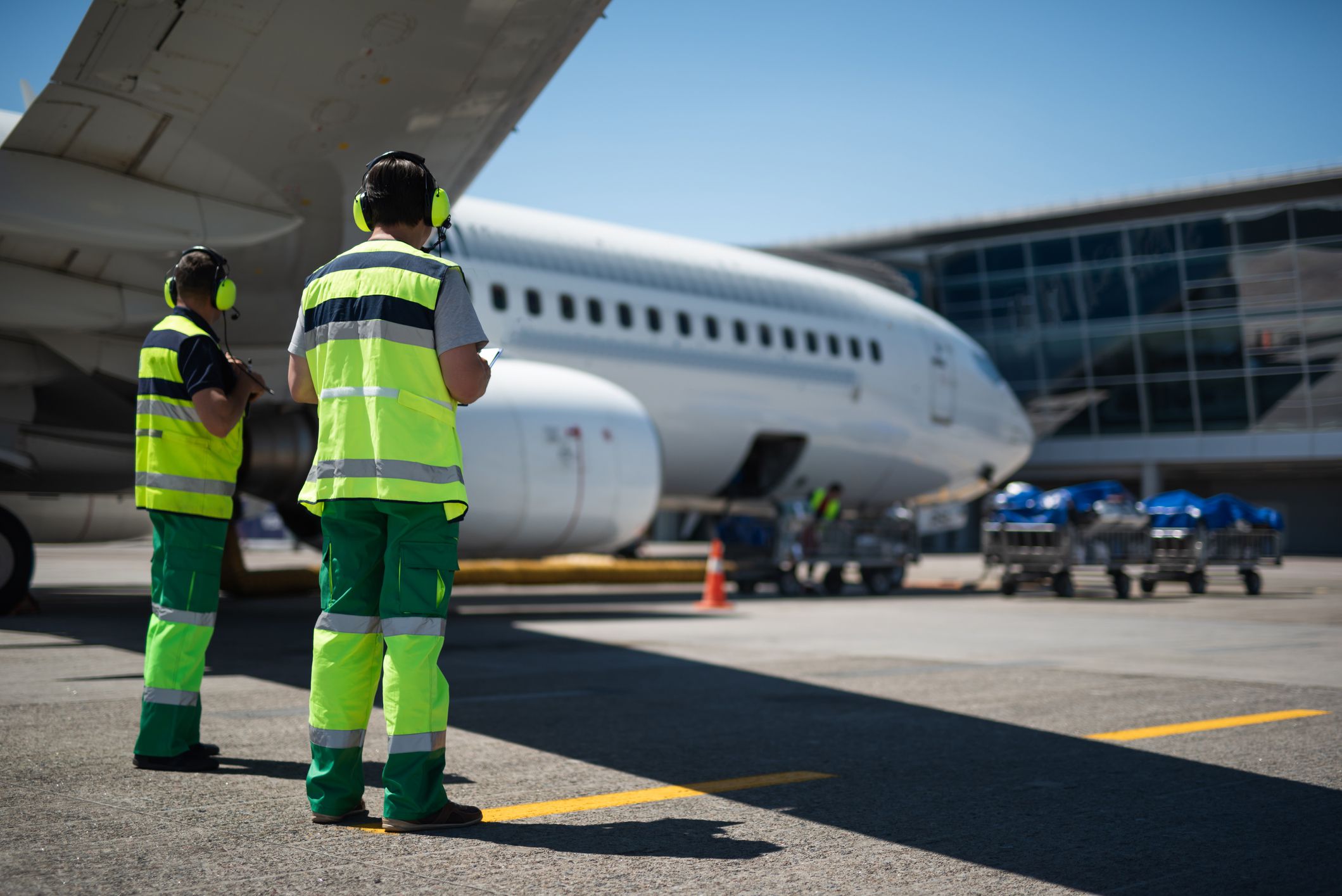 Two aviation crewmembers on the tarmac on the left foreground with a large passenger airplane with luggage trolleys blurred in the background, at a gate in an airport