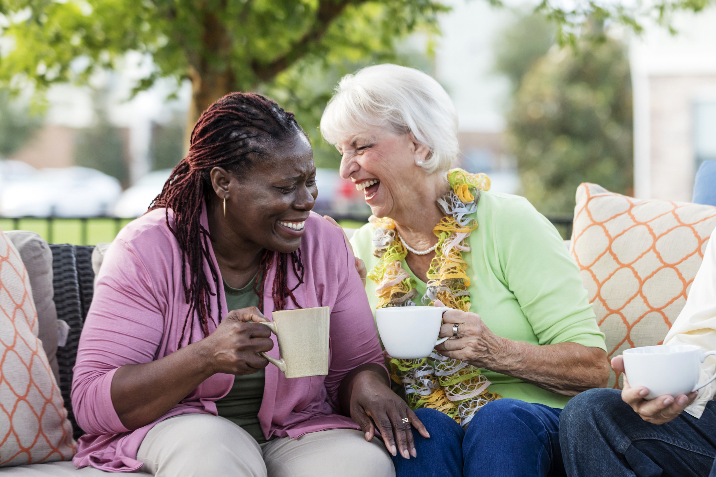 Two female senior friends laughing together while each holding a mug, on an patio sofa outside, with another senior man sitting on the right side, a large tree and apartment building blurred in the background